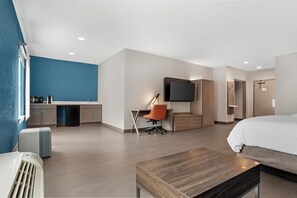 A modern hotel room with a kitchenette, a TV mounted on the wall, a bed, and a desk with an orange chair.