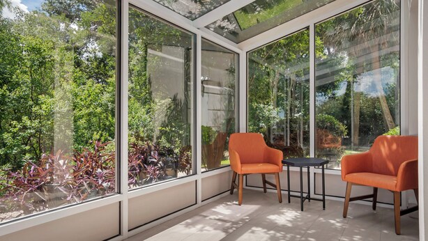 A conservatory with large glass windows, two orange chairs, a small table, and a view of greenery outside.