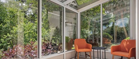 A conservatory with large glass windows, two orange chairs, a small table, and a view of greenery outside.