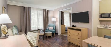 A hotel room with a wooden floor, a desk, a chair, a sofa, a television, a dresser, and a mirror.