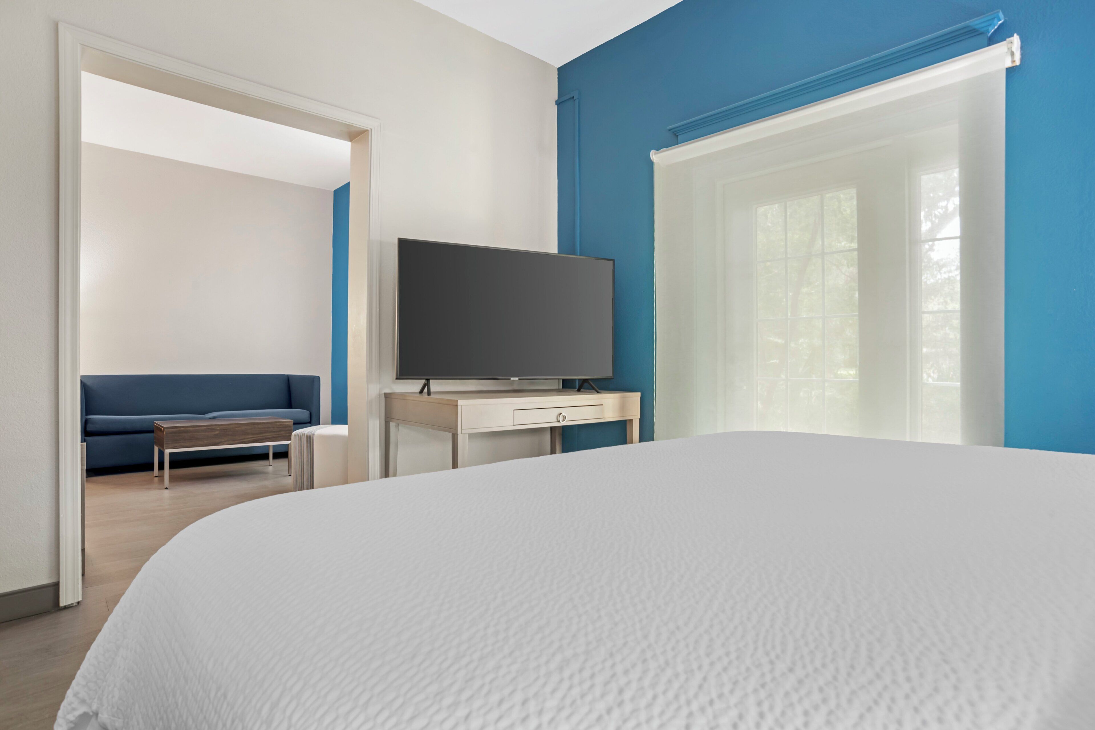 A hotel room with a large bed, a TV on a bedside table, a sofa, and a window with blinds.