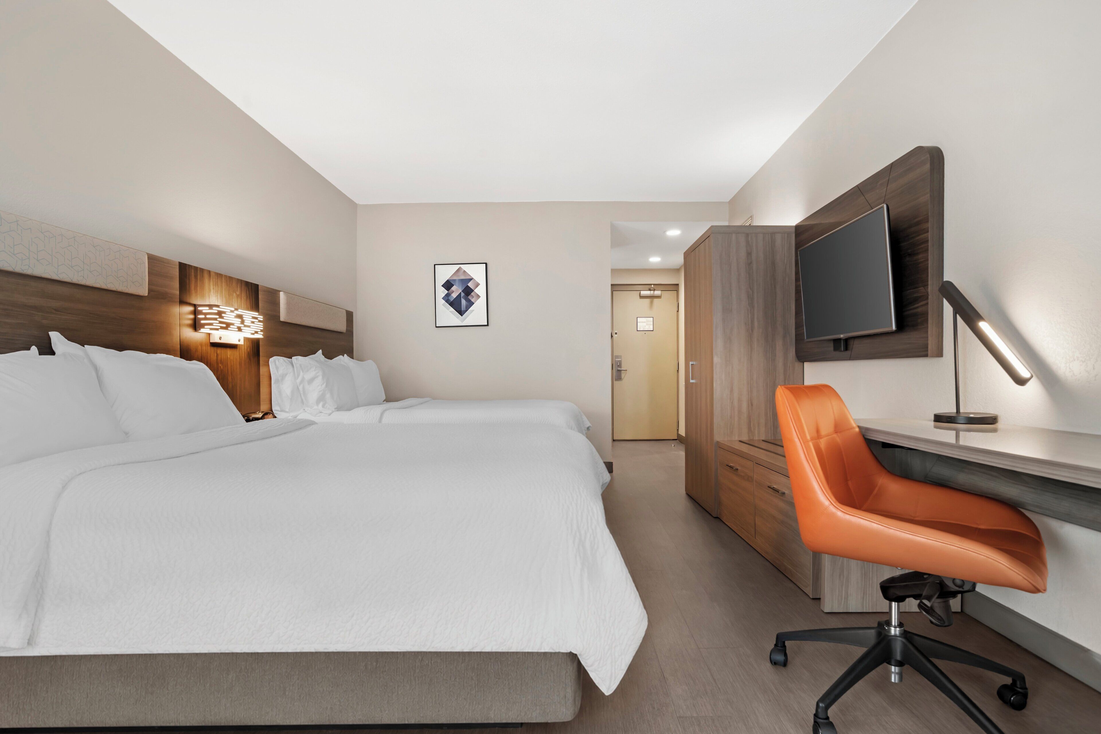A hotel room with a large bed, a desk with a chair, a TV mounted on the wall, and a framed picture on the wall.