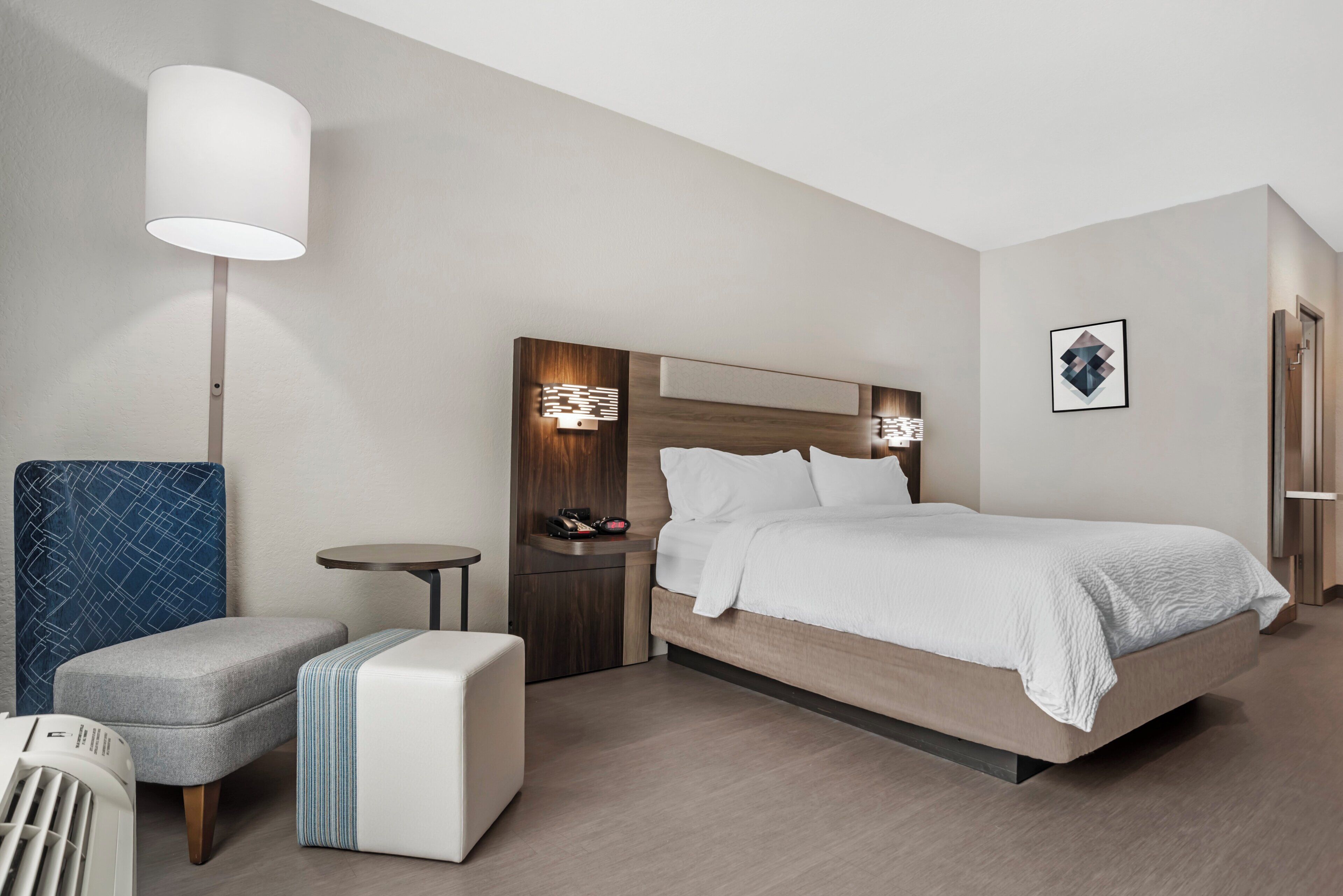 A hotel room with a bed, bedside table, lamp, chair, and a small table.