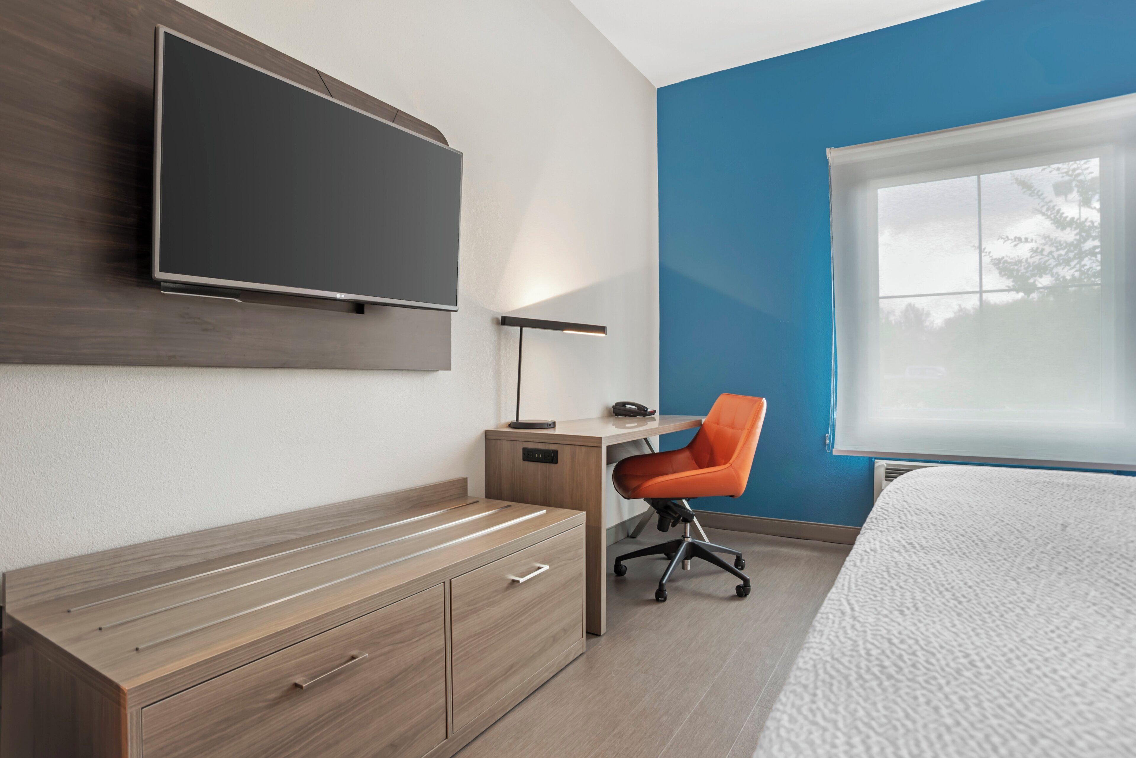 A modern bedroom with a bed, a desk with a chair, a television, and a window.