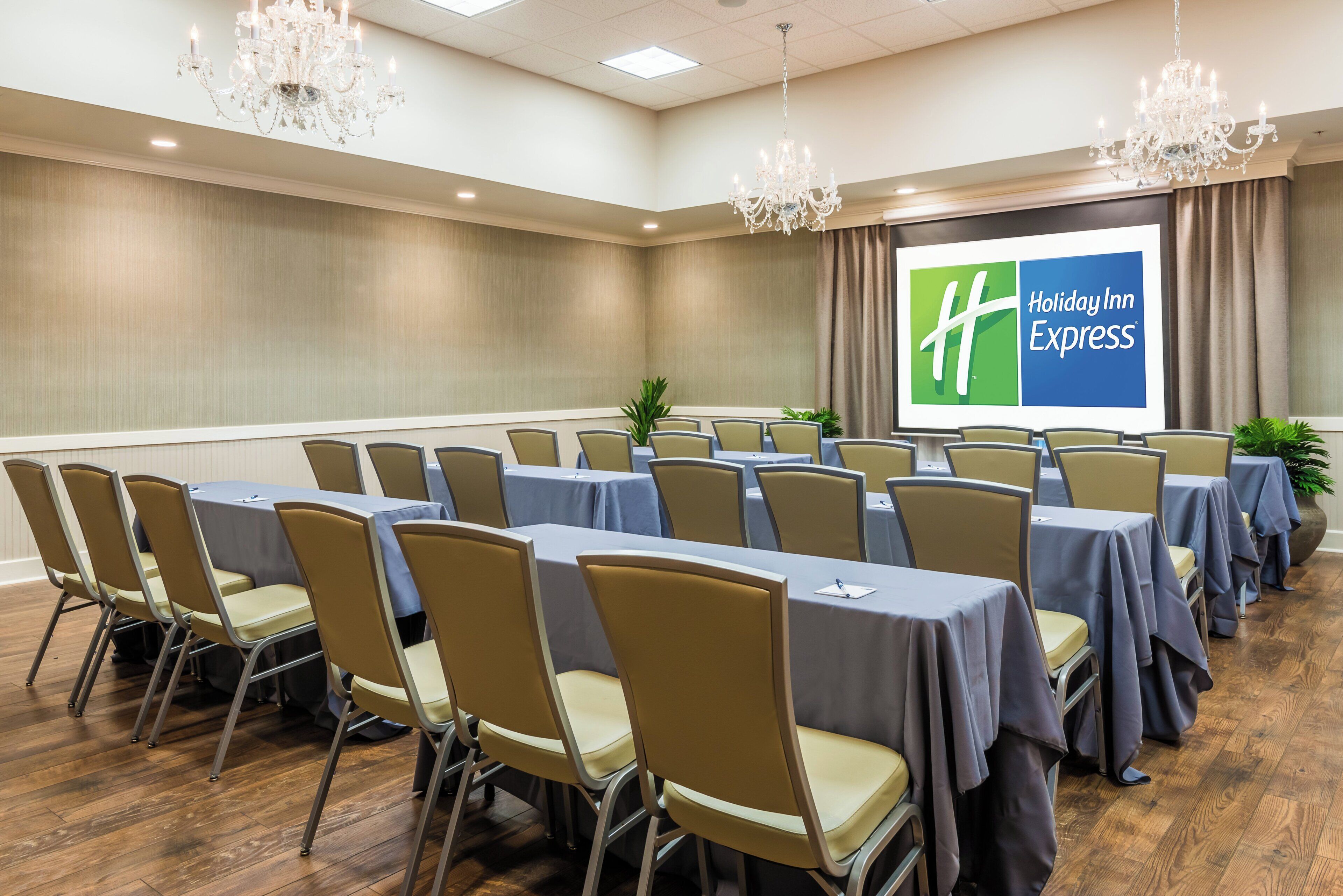 A conference room with rows of tables and chairs, a projector screen with the Holiday Inn Express logo, and chandeliers.