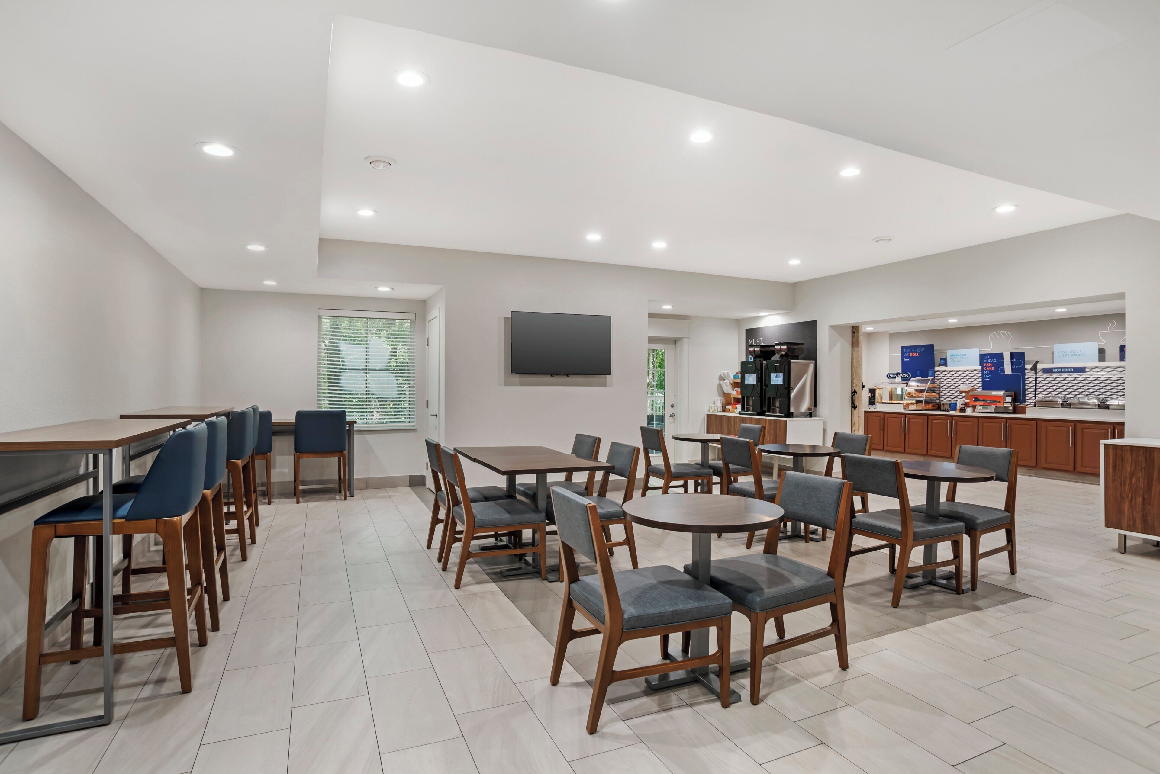 A spacious dining area with multiple tables and chairs, a flat-screen TV mounted on the wall, and a kitchen area in the background.