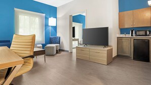A modern hotel room with a flat-screen TV, a dining area with a table and chairs, a kitchenette, and a large window.