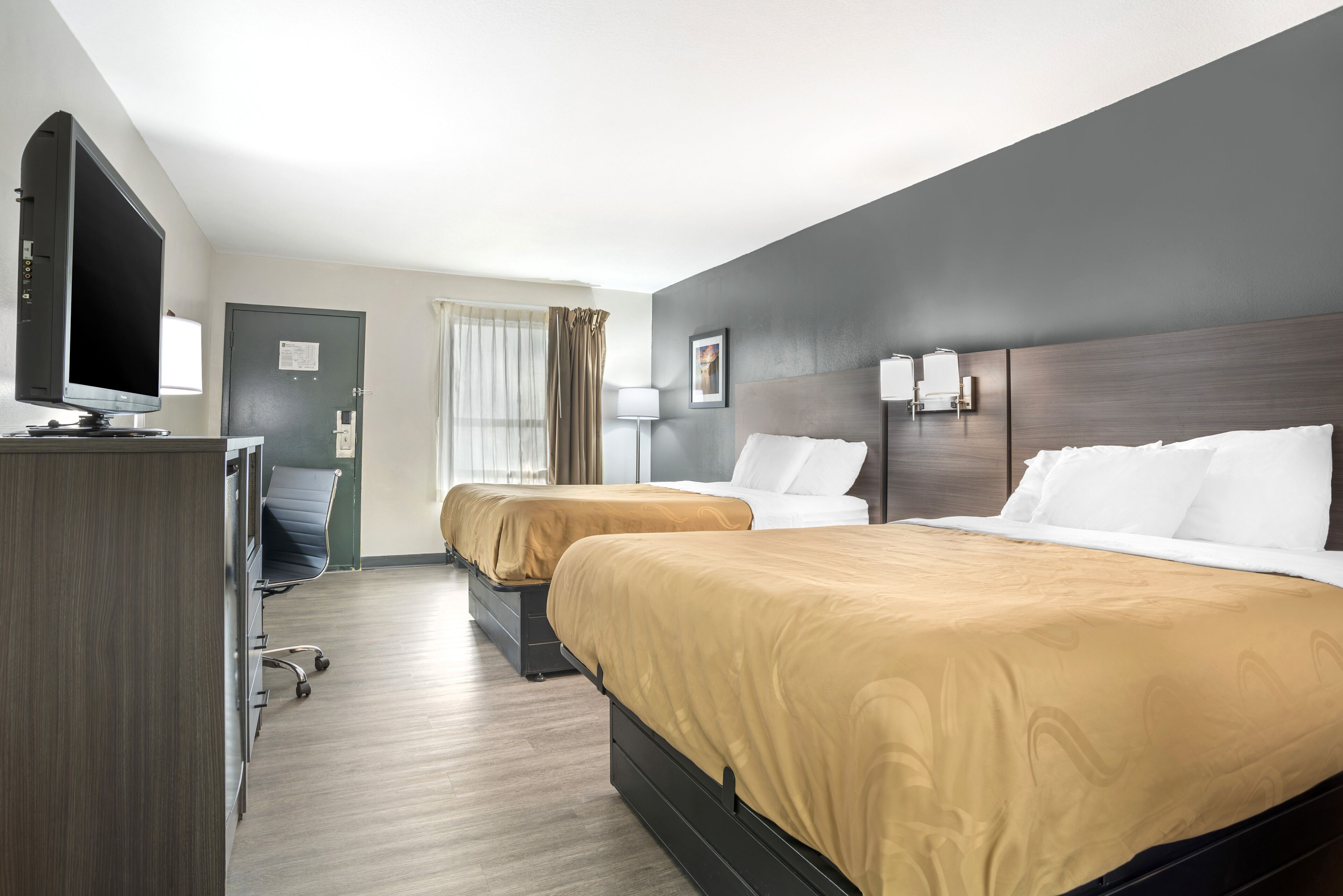 A hotel room with two beds, a desk with a flat-screen TV, a chair, and a door.
