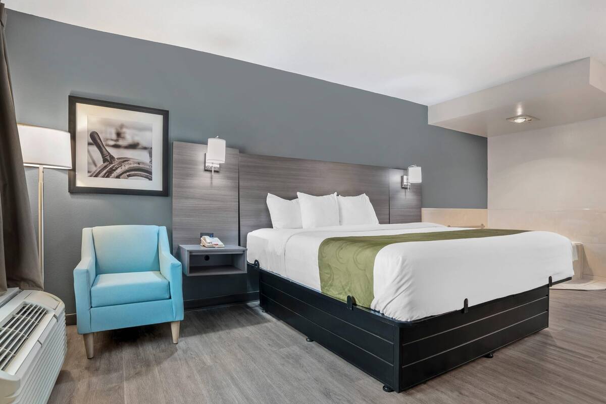 Una camera d'albergo con un letto, un comodino, una sedia, un quadro incorniciato e una lampada a parete.