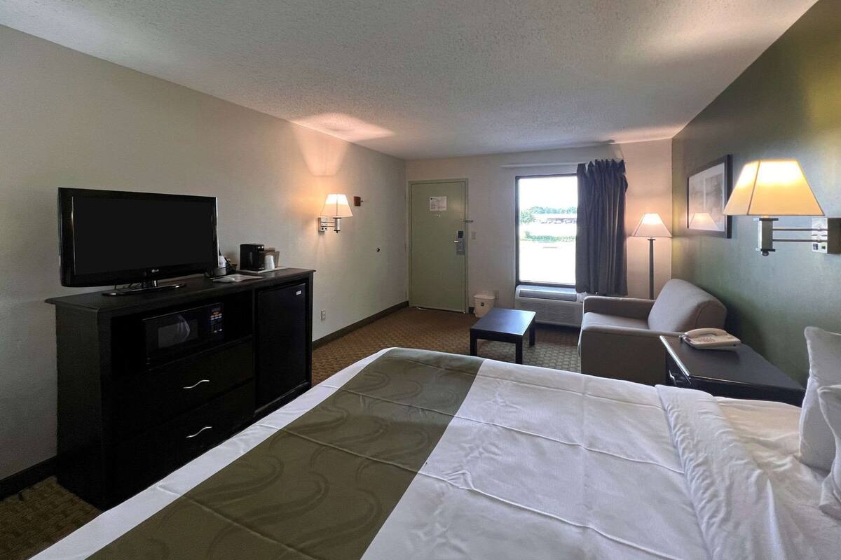 Una camera d'albergo con un letto, una TV, una scrivania, una sedia, un divano e una finestra con tende.