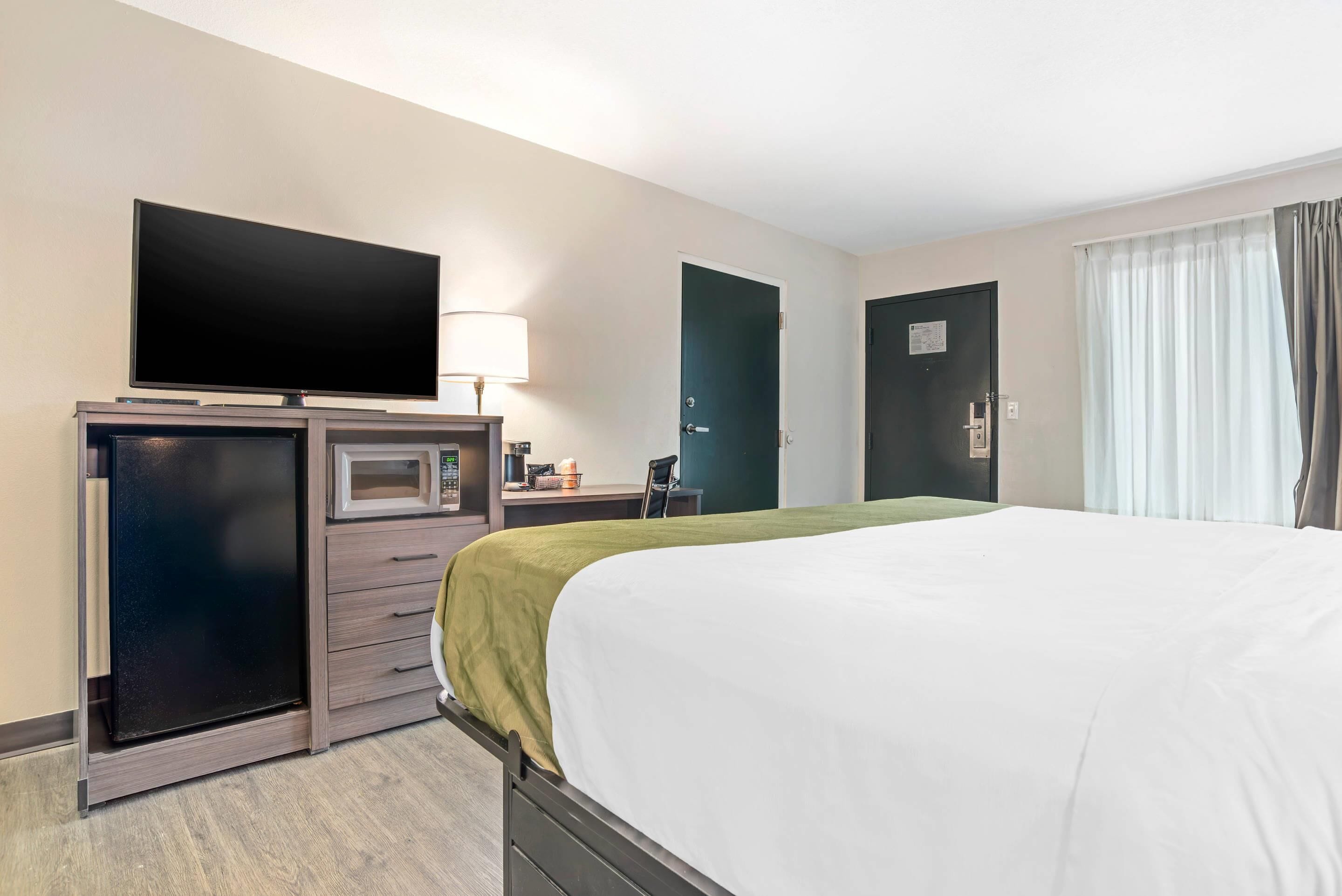 A hotel room with a bed, a flat-screen TV, a microwave, a desk with a lamp, and a door.