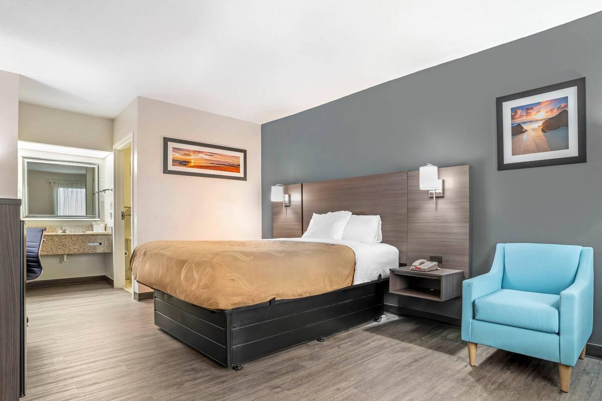 Una camera d'hotel con un letto, una sedia, un comodino, uno specchio e opere d'arte alle pareti.