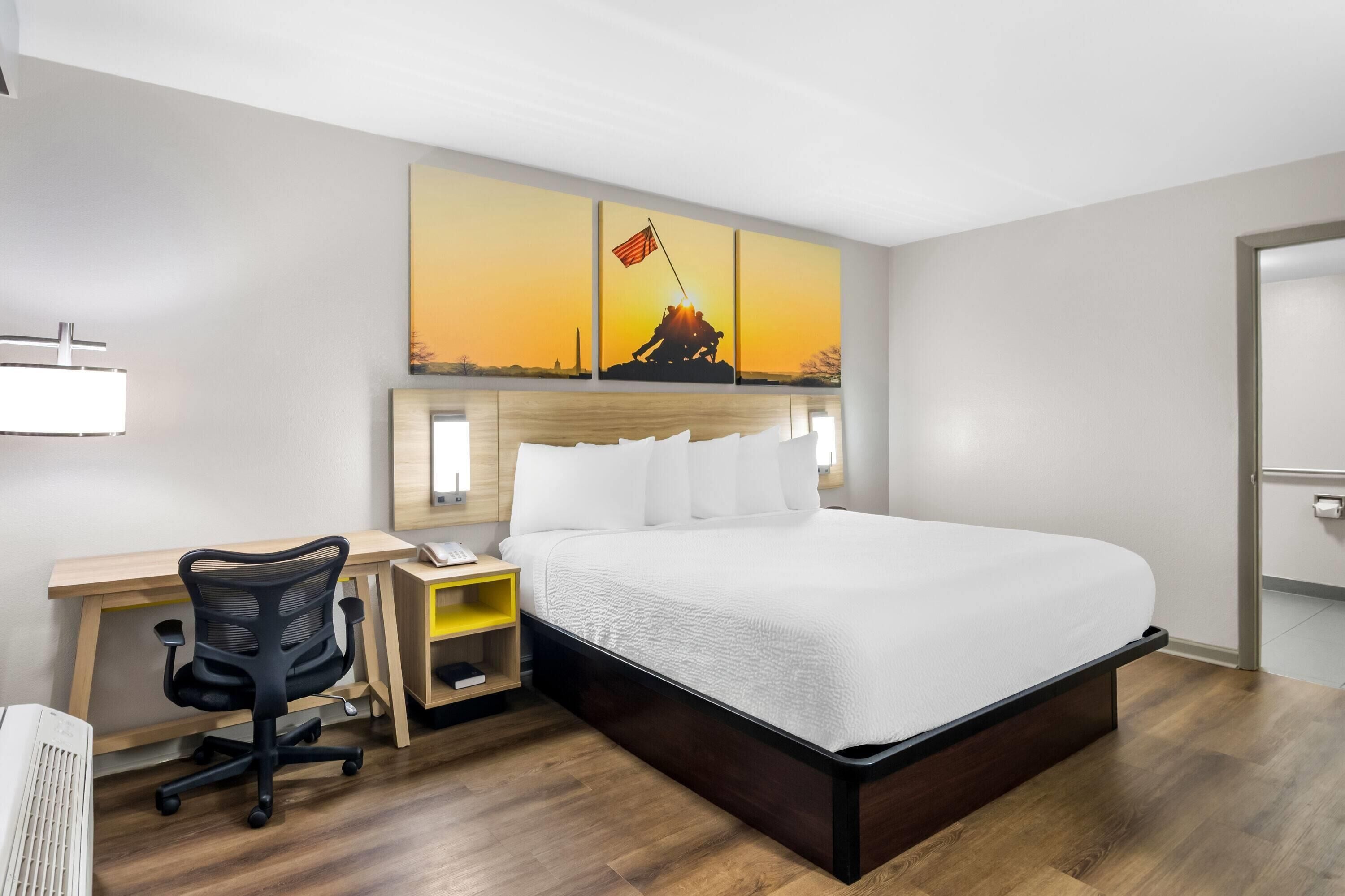 A hotel room with a bed, desk, chair, and a painting of a sunset.