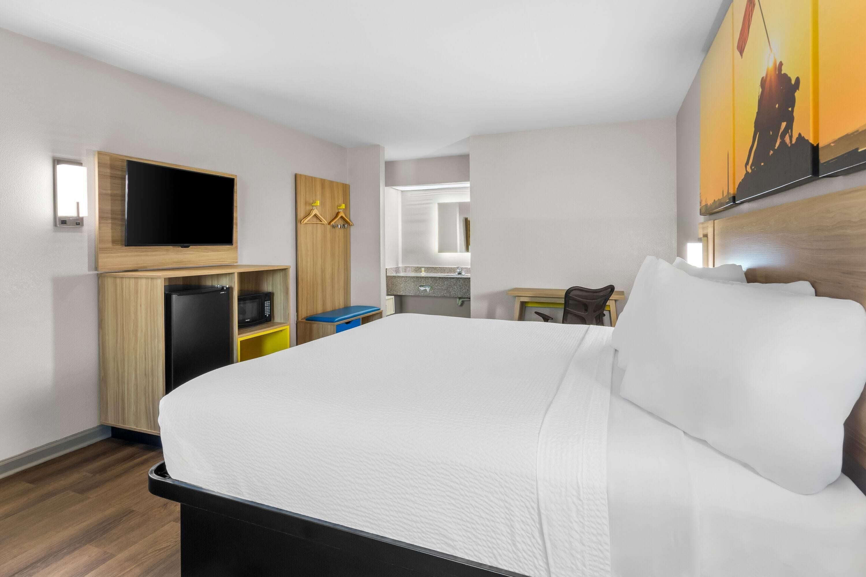 Habitación de hotel con una cama grande, un televisor de pantalla plana montado en la pared, una pequeña kitchenette y un baño visible a través de una puerta abierta.