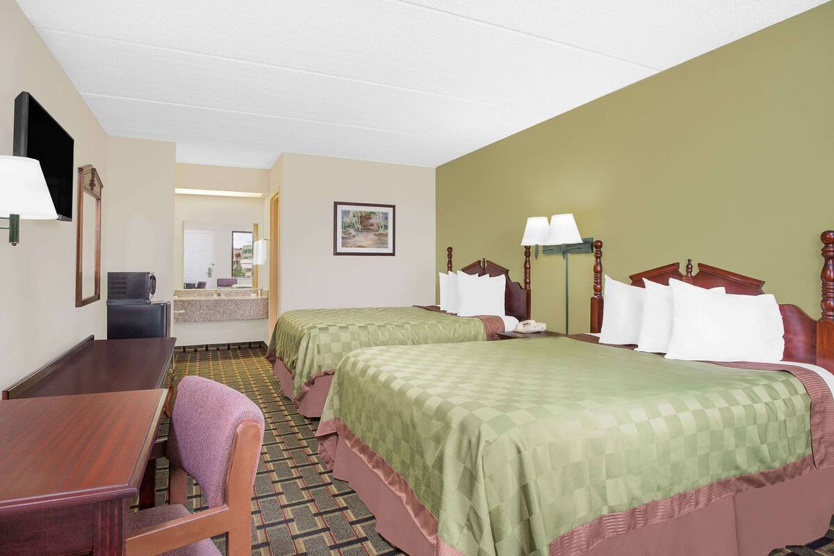 Quarto de hotel com duas camas, uma escrivaninha, uma cadeira, televisão e banheiro visível por uma porta aberta.