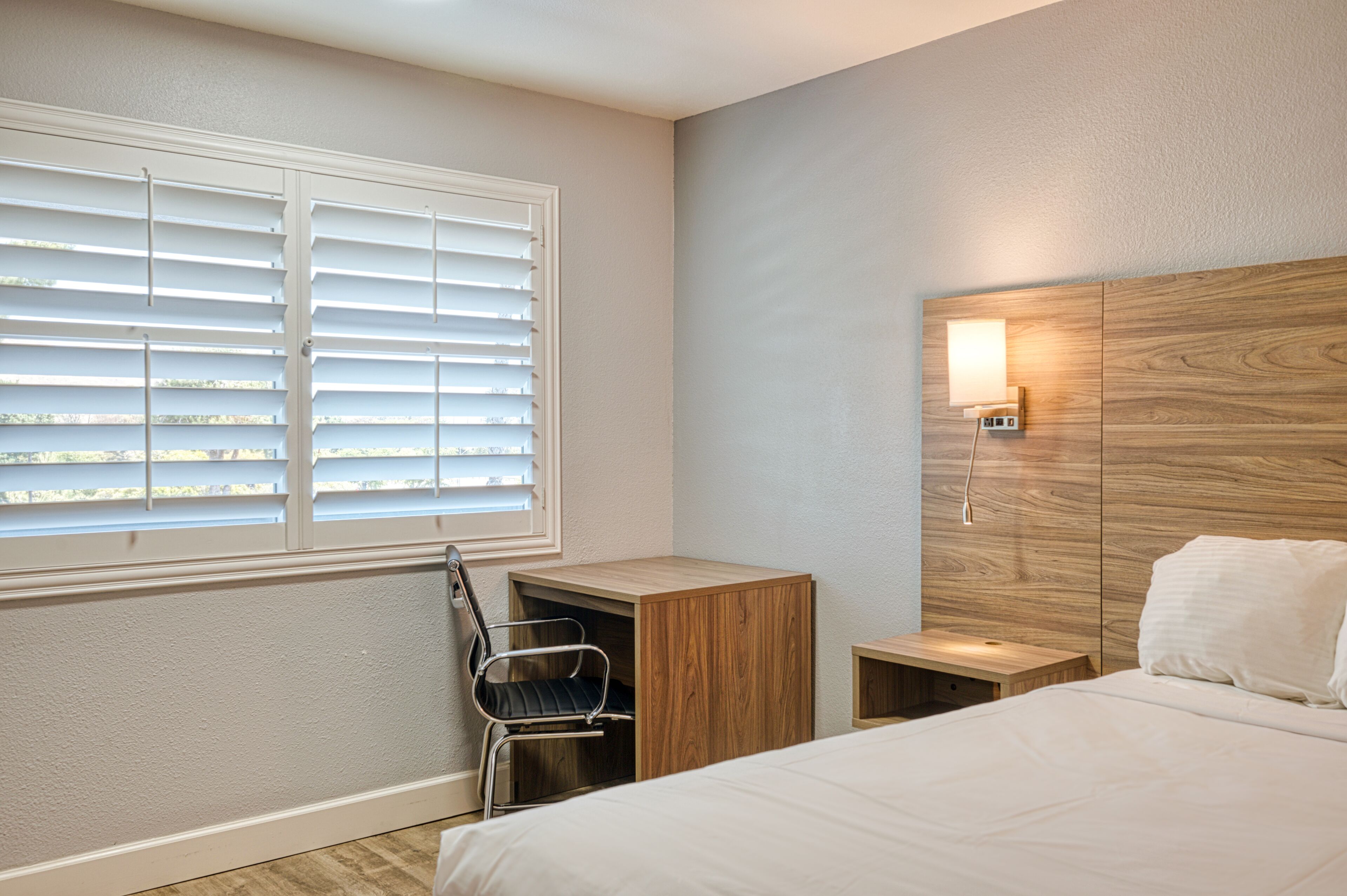 Une chambre d’hôtel comprenant un lit, un bureau avec une chaise, une table de chevet, une lampe fixée au mur et une fenêtre avec des stores.