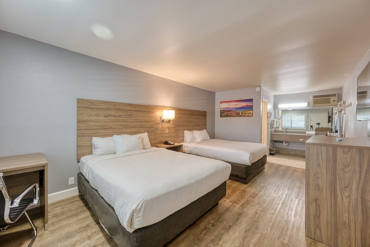 Une chambre d’hôtel avec deux lits, une tête de lit en bois, une table de chevet avec une lampe, un bureau avec une chaise et une salle de bain avec un miroir et un lavabo.