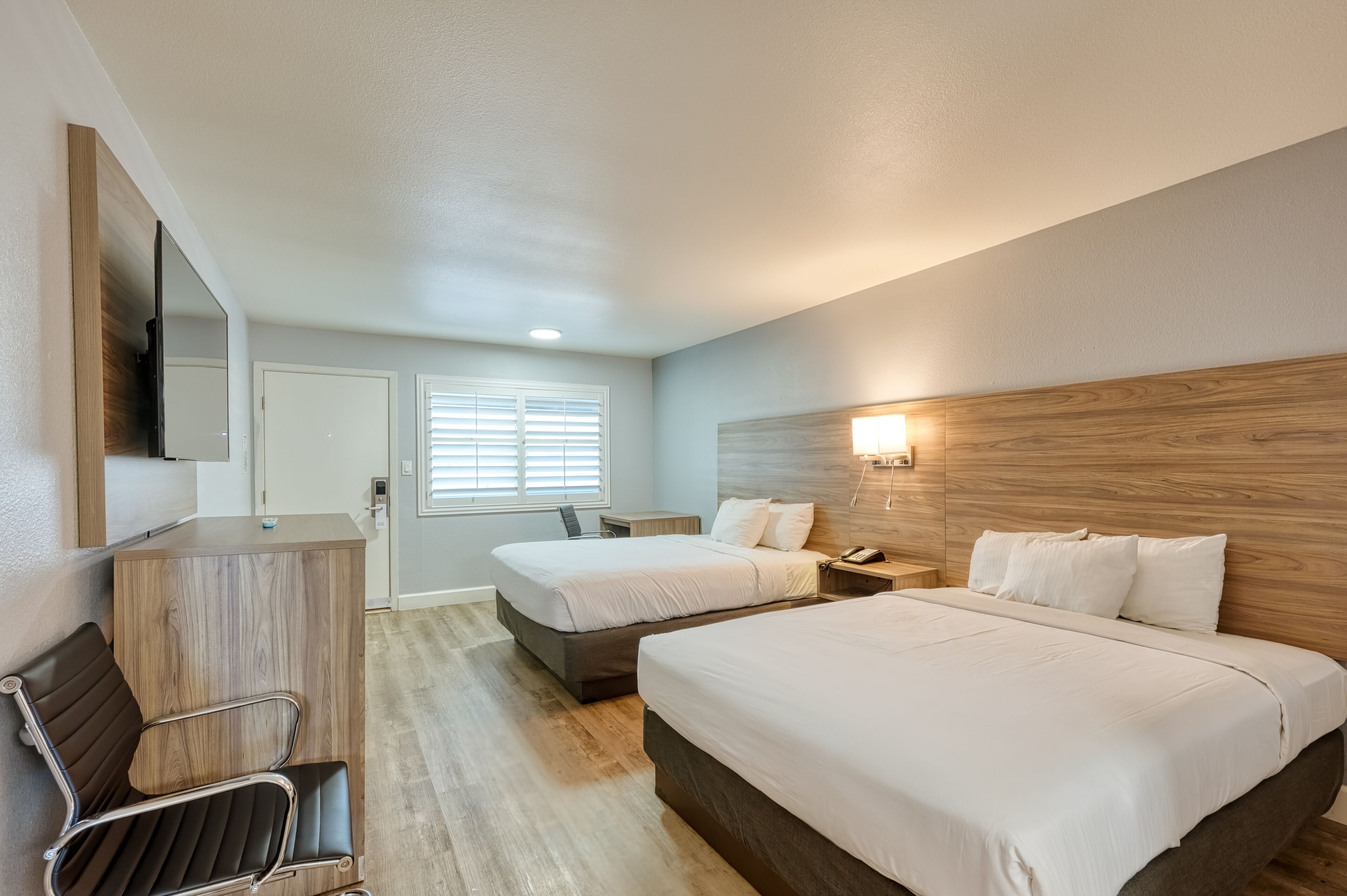 A hotel room with two beds, a desk, a chair, a mirror, and a window with blinds.