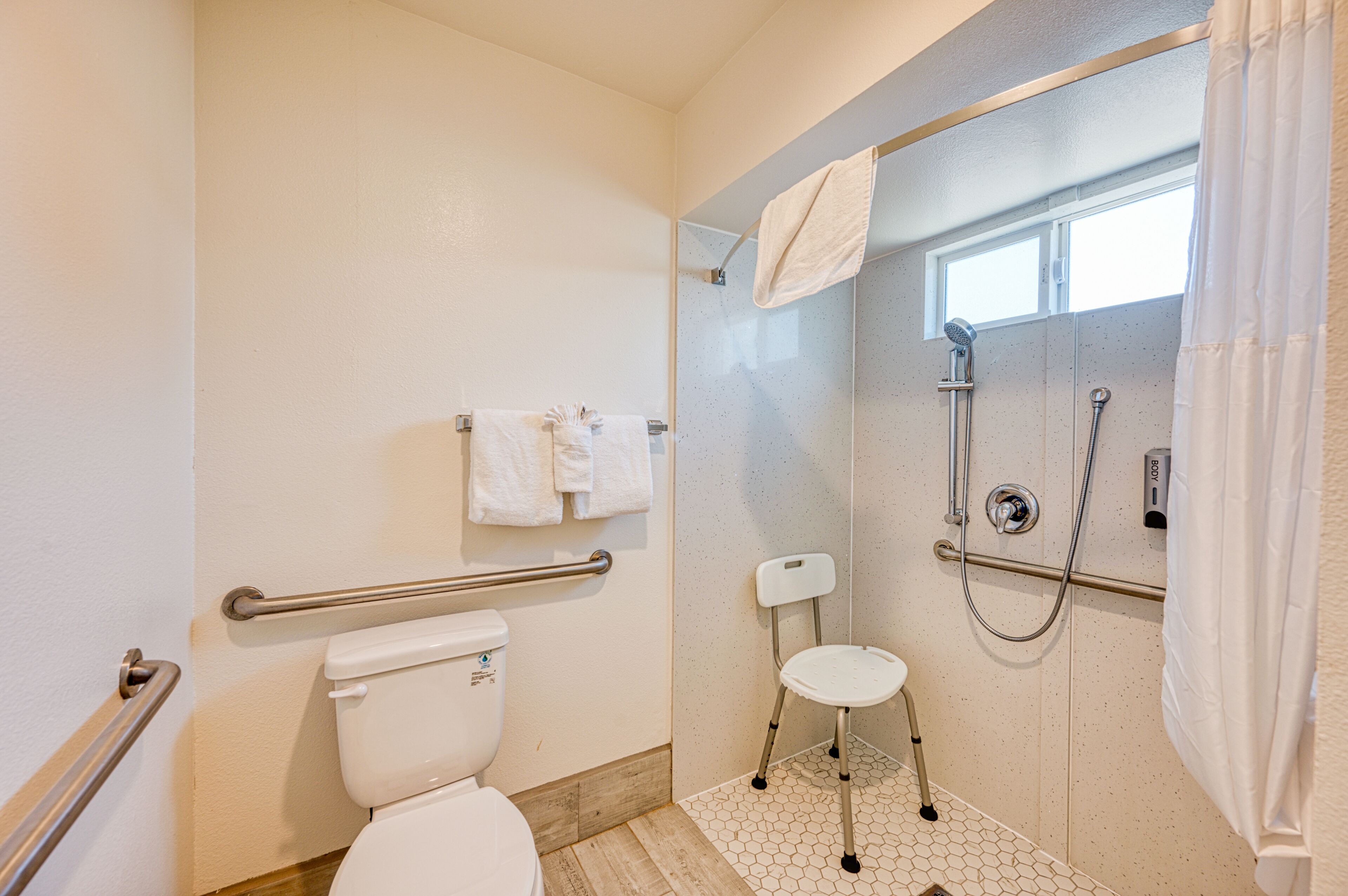 A bathroom with a toilet, a shower with a glass door, a grab bar, and a white chair.