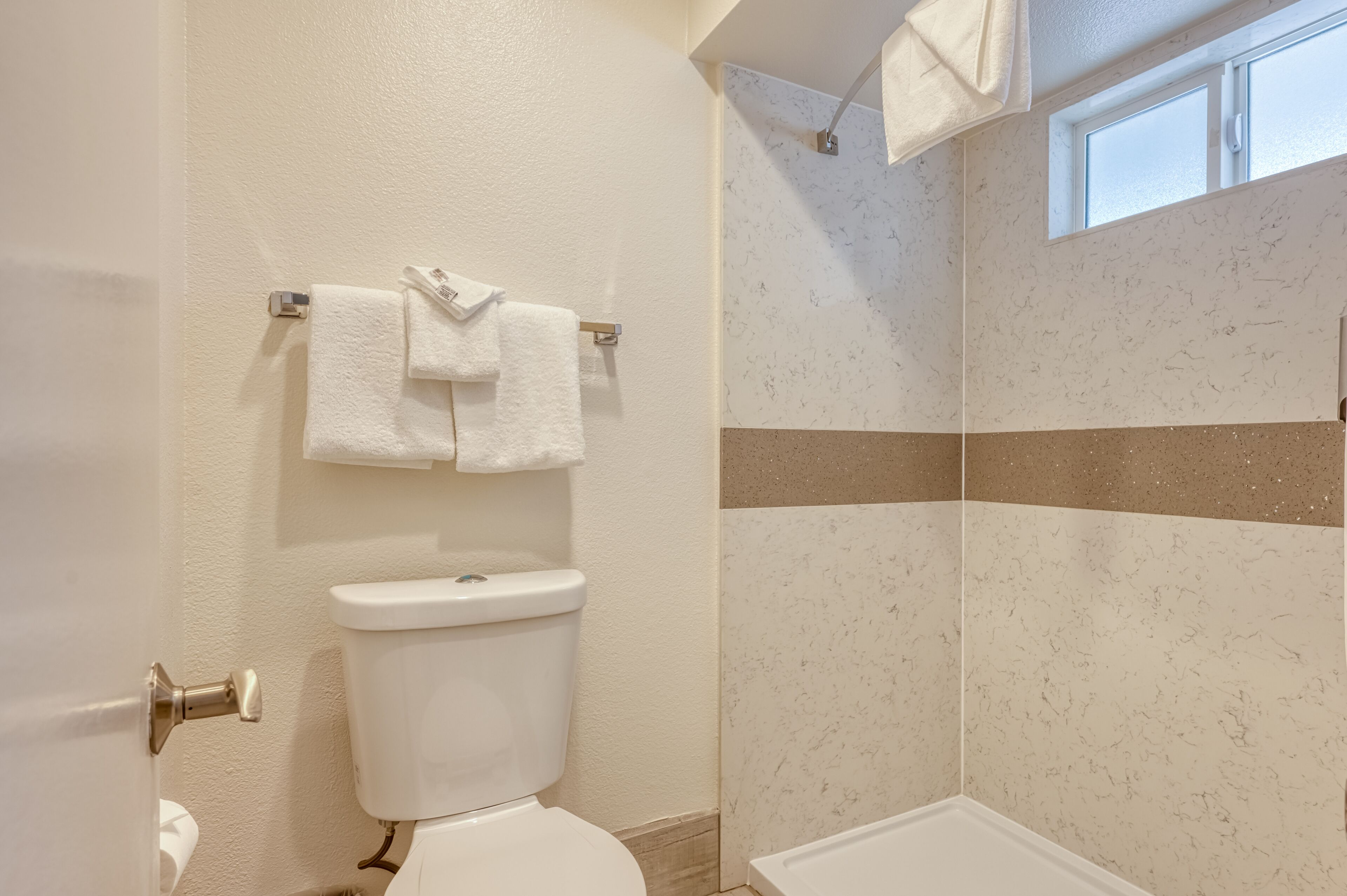 A bathroom with a toilet, a towel rack with two towels, a shower with a beige and white tiled wall, and a window above the shower.
