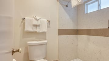 A bathroom with a toilet, a towel rack with two towels, a shower with a beige and white tiled wall, and a window above the shower.