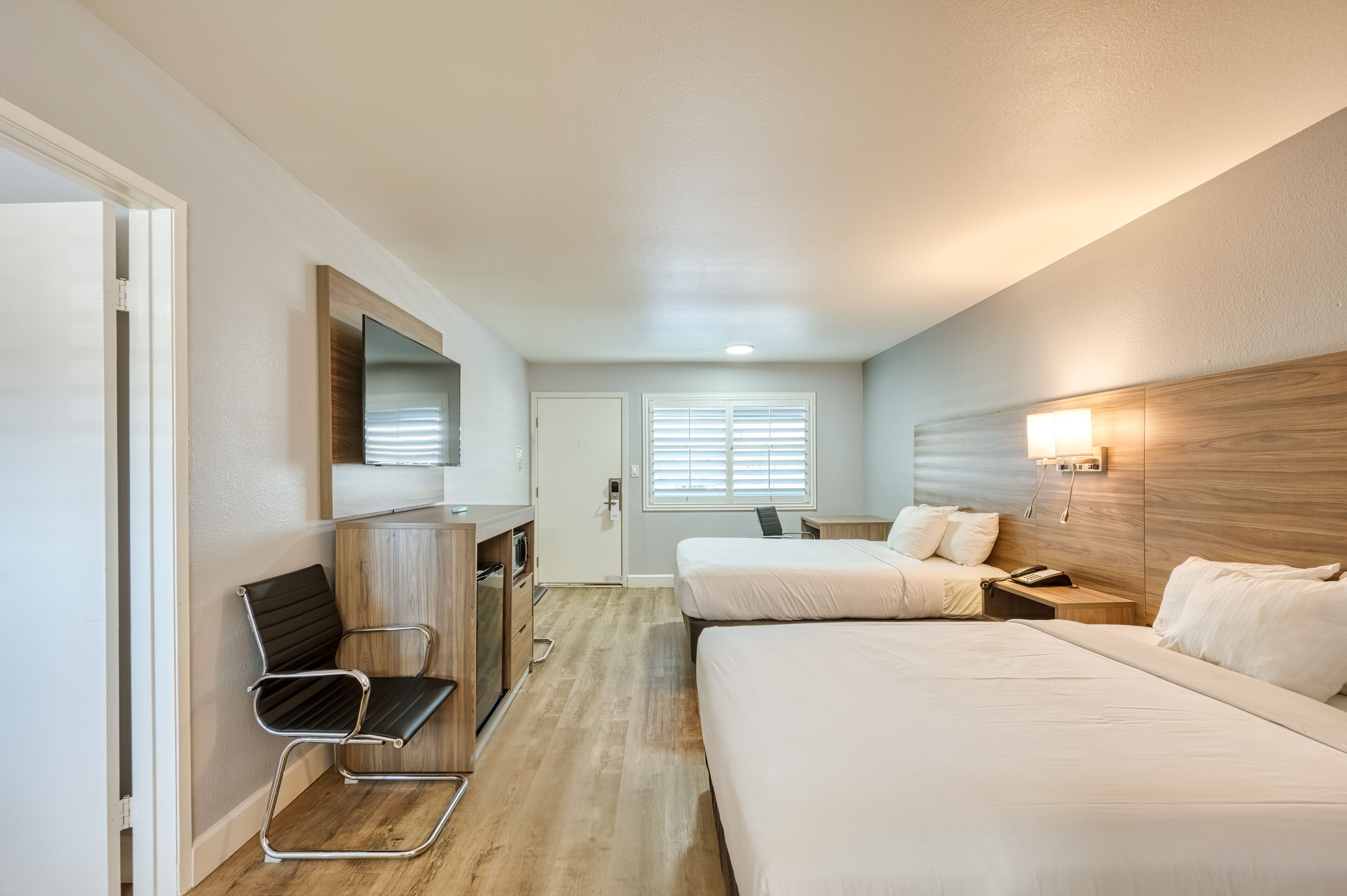 Habitación de hotel con dos camas, un escritorio con silla, un televisor y una ventana con persianas.