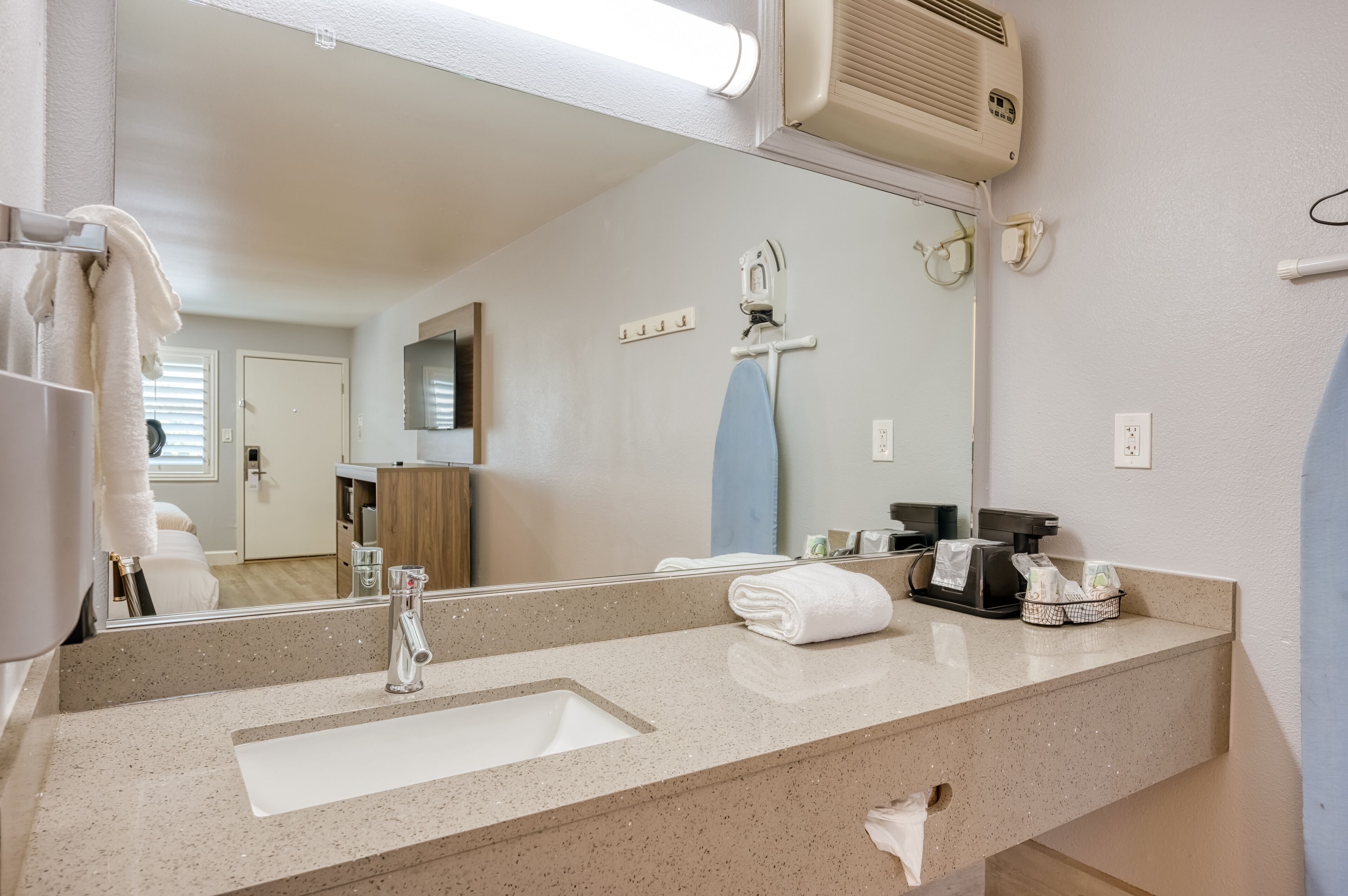 A hotel bathroom with a large mirror, a sink, a towel rack, and a towel dispenser.