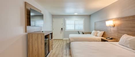 A hotel room with two beds, a TV mounted on the wall, a desk with a lamp, and a window with blinds.