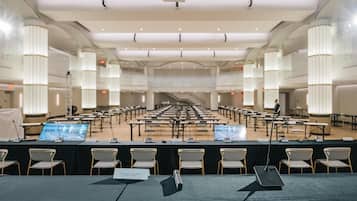 Un amplio salón de conferencias con filas de mesas y sillas, un escenario con un atril, y una persona de pie cerca del escenario.