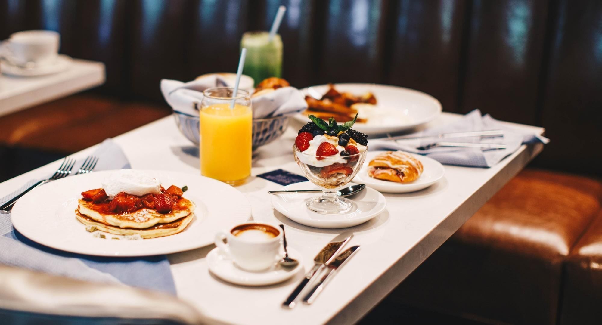 Een ontbijttafel met pannenkoeken, een fruitparfait, een croissant en een glas sinaasappelsap.