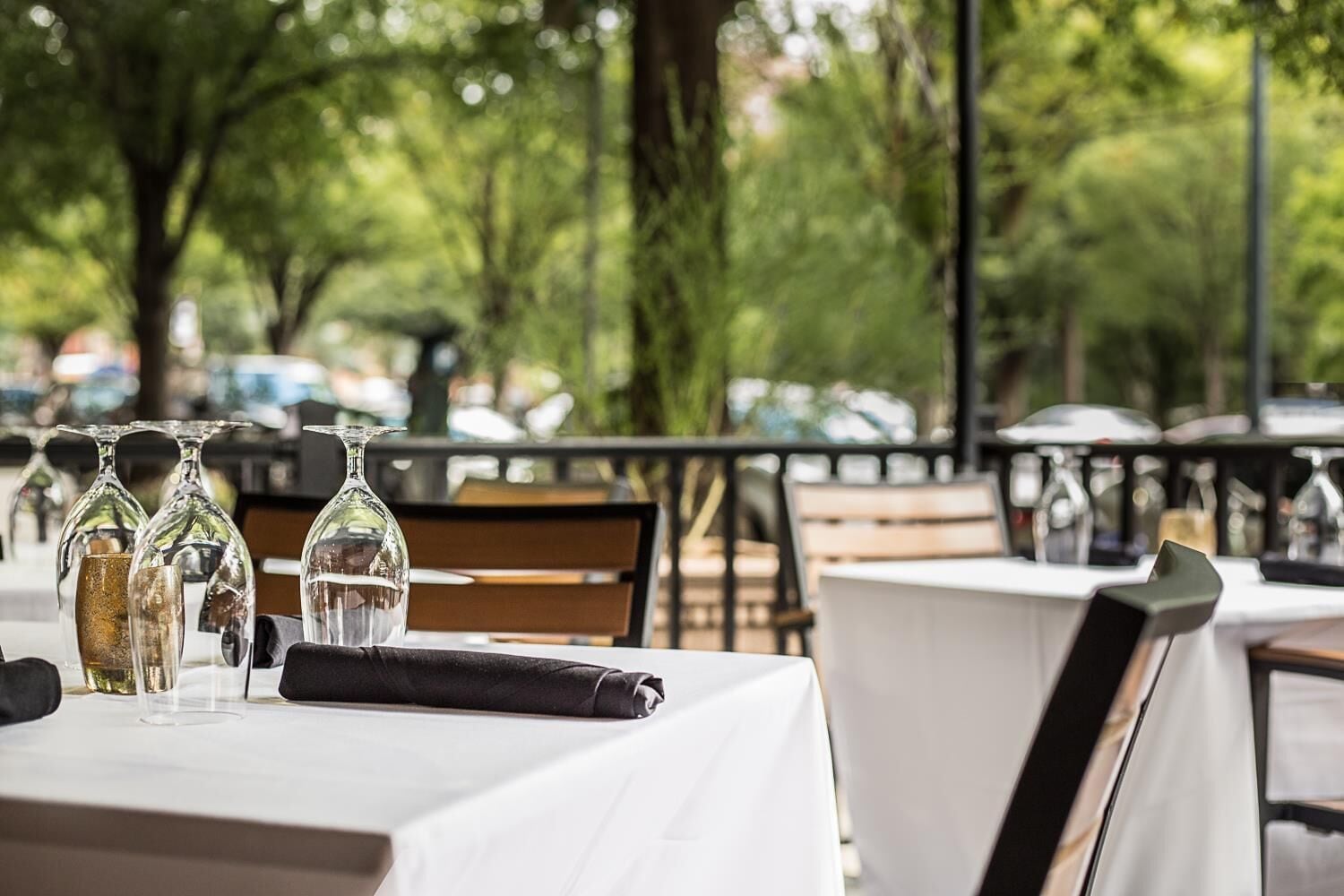 An outdoor dining area with tables set for two, each with a white tablecloth, clear glassware, and a folded napkin.