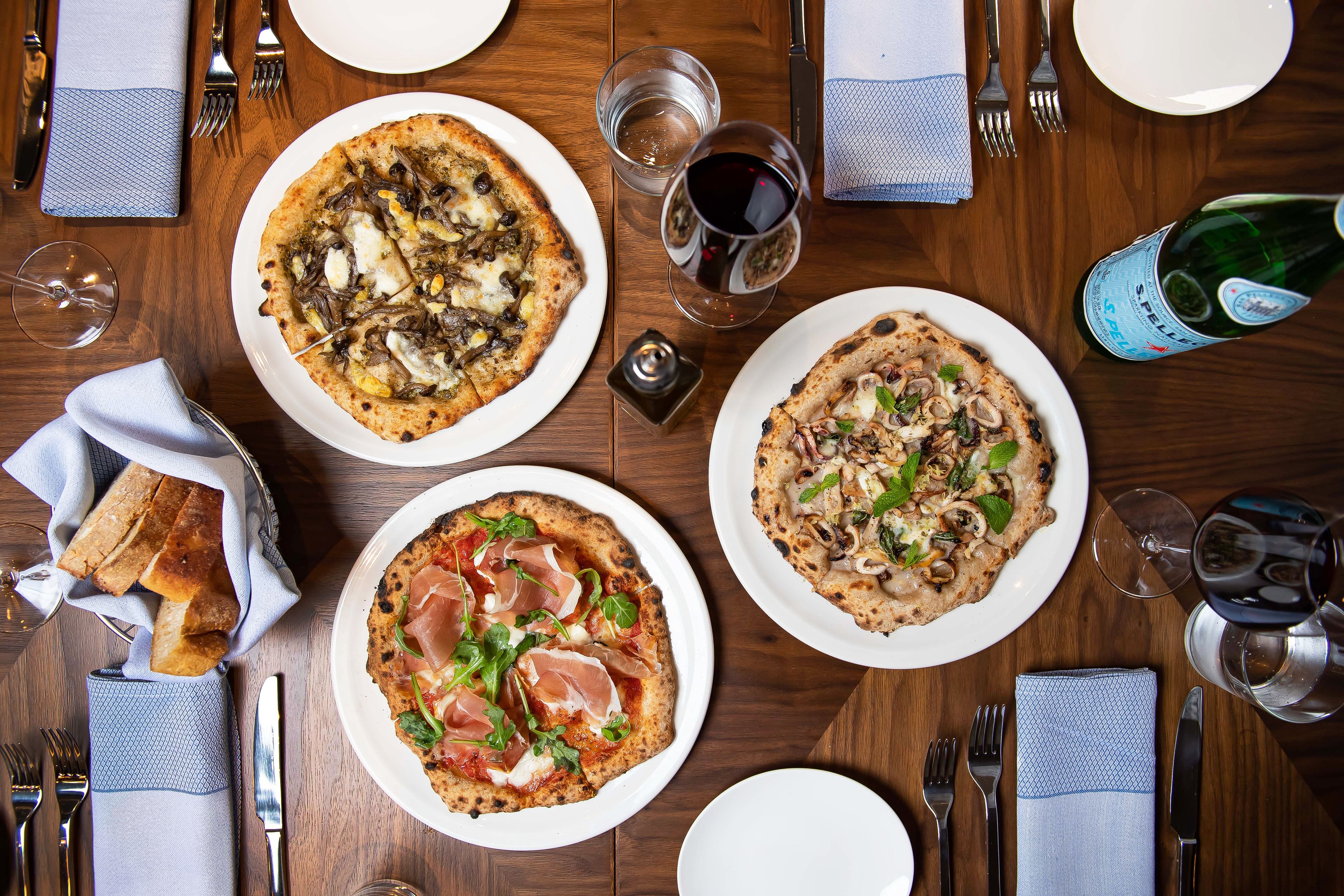 Une table à manger avec trois pizzas, des verres à vin et une bouteille de bière Stella Artois.