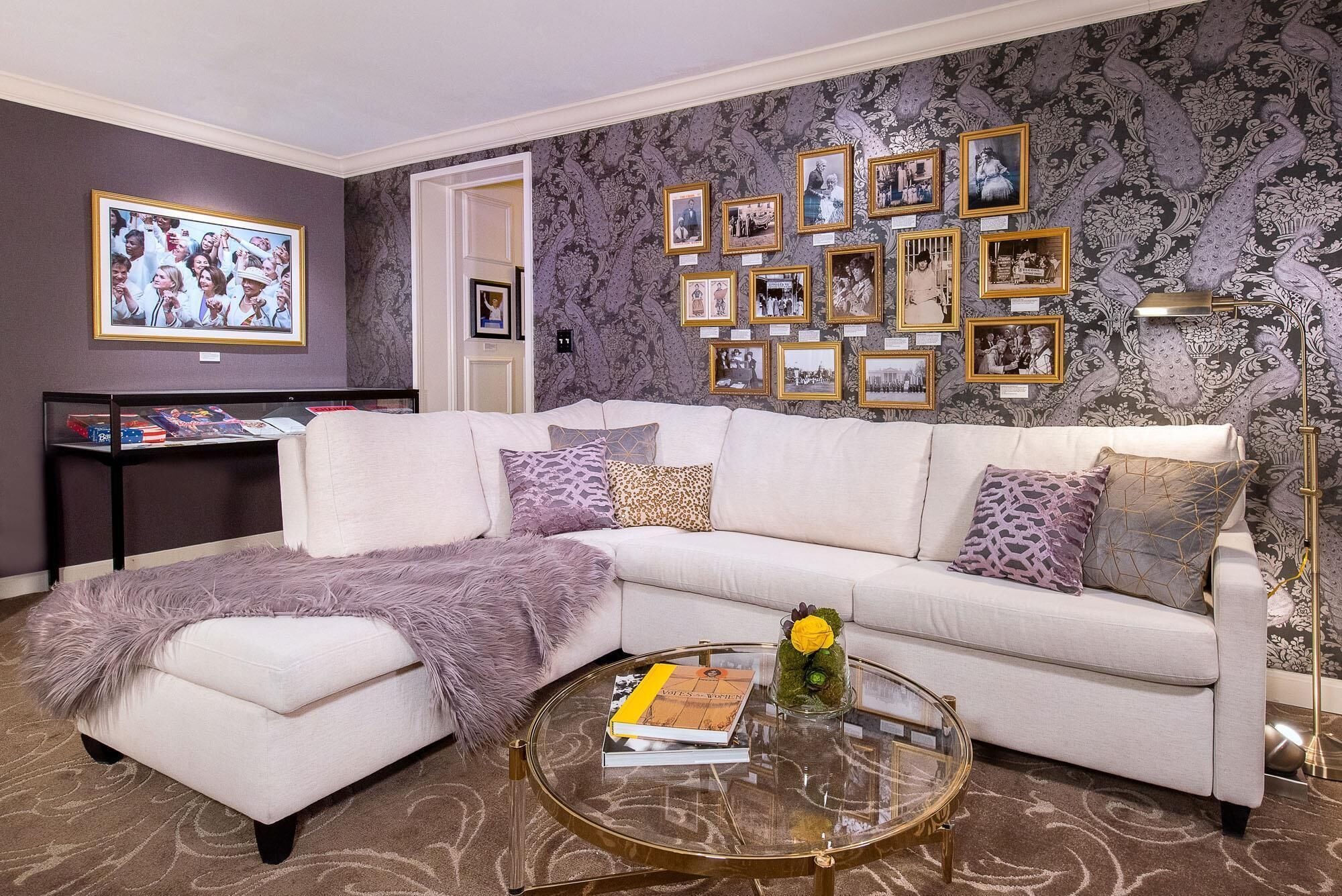 A living room with a white sofa, multiple framed pictures on the wall, a glass coffee table with a book, and a decorative vase.