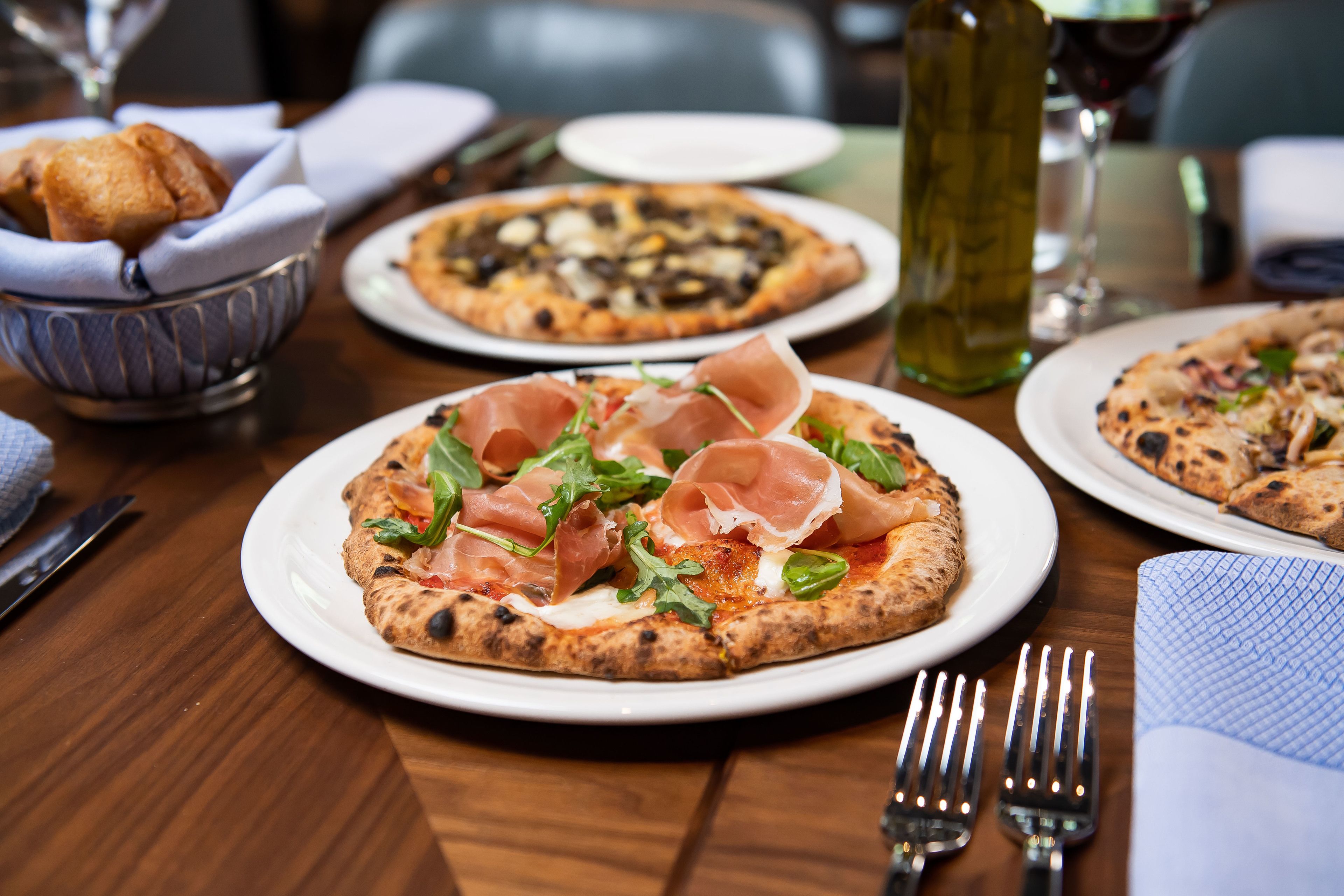 Una mesa de comedor con pizzas, una botella de aceite de oliva y cubiertos.