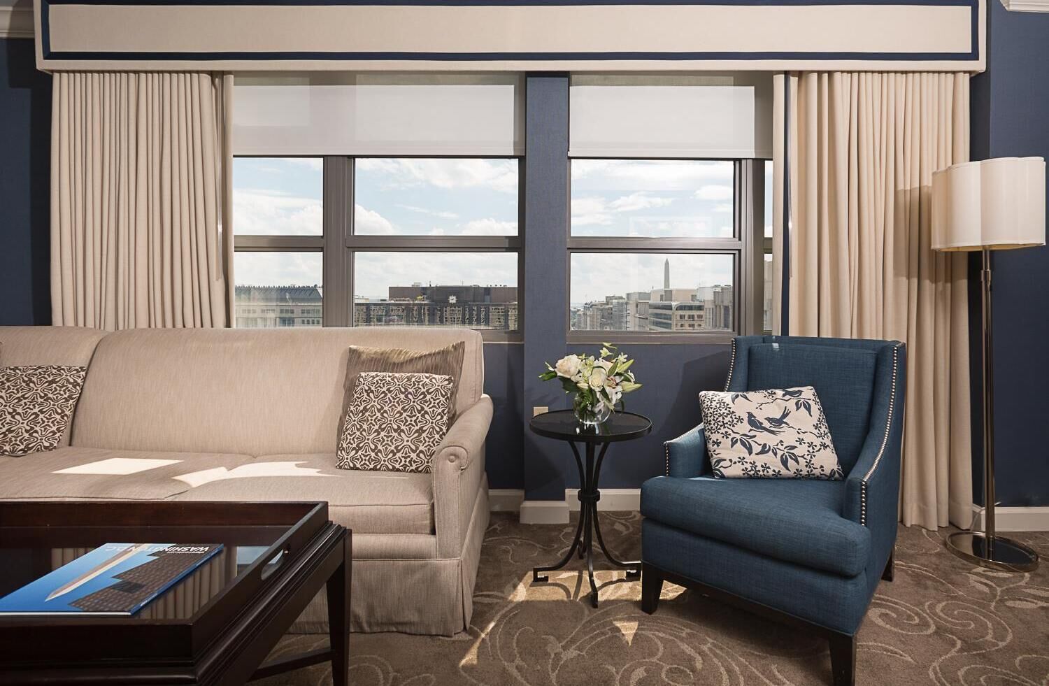 A hotel room with a beige sofa, a blue armchair, a small round table with a vase of flowers, and a floor lamp.