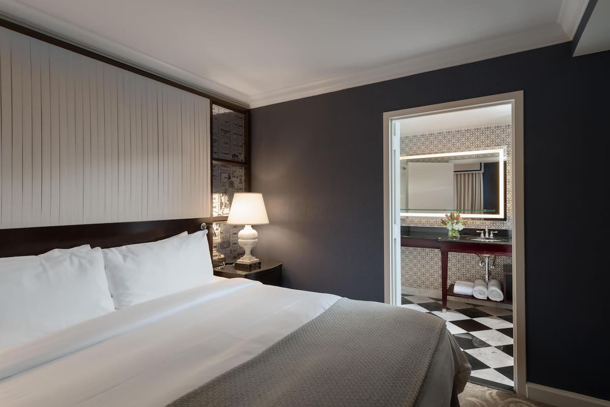 A hotel room with a large bed, a nightstand with a lamp, a dark wall, a patterned mirror, and a checkered floor.