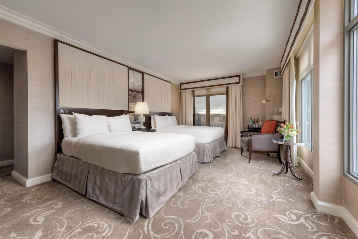 A hotel room with two beds, a small table with a lamp, a chair, and a view of the city through large windows.