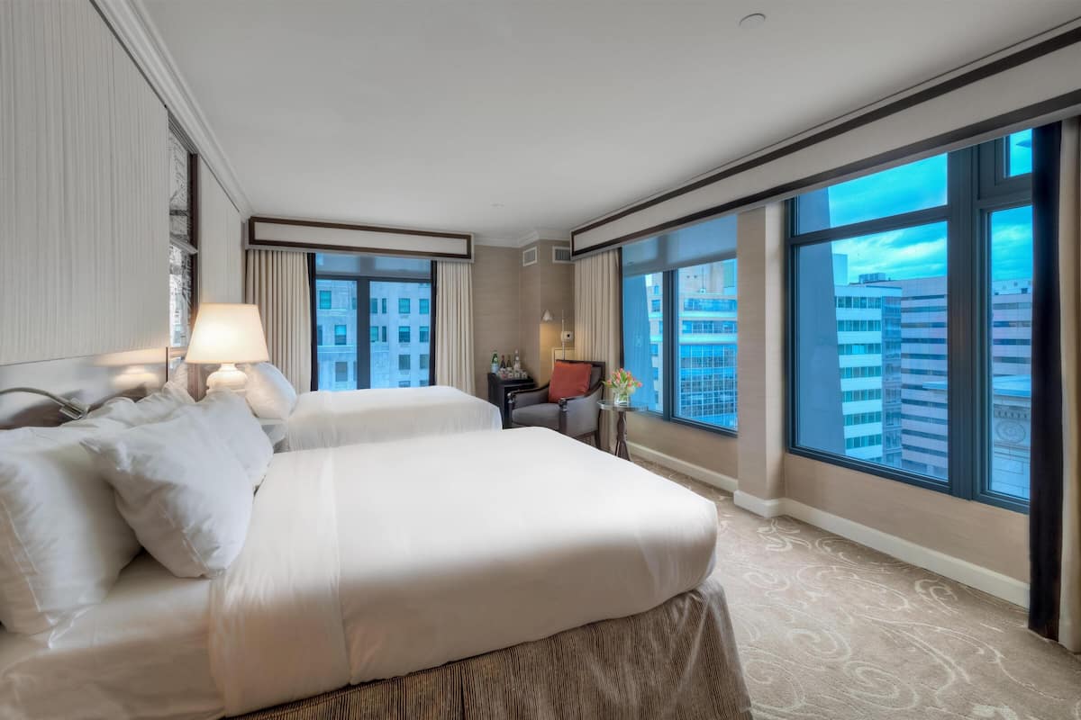 A hotel room with two beds, a large window with a city view, a small table with a lamp, and a chair.