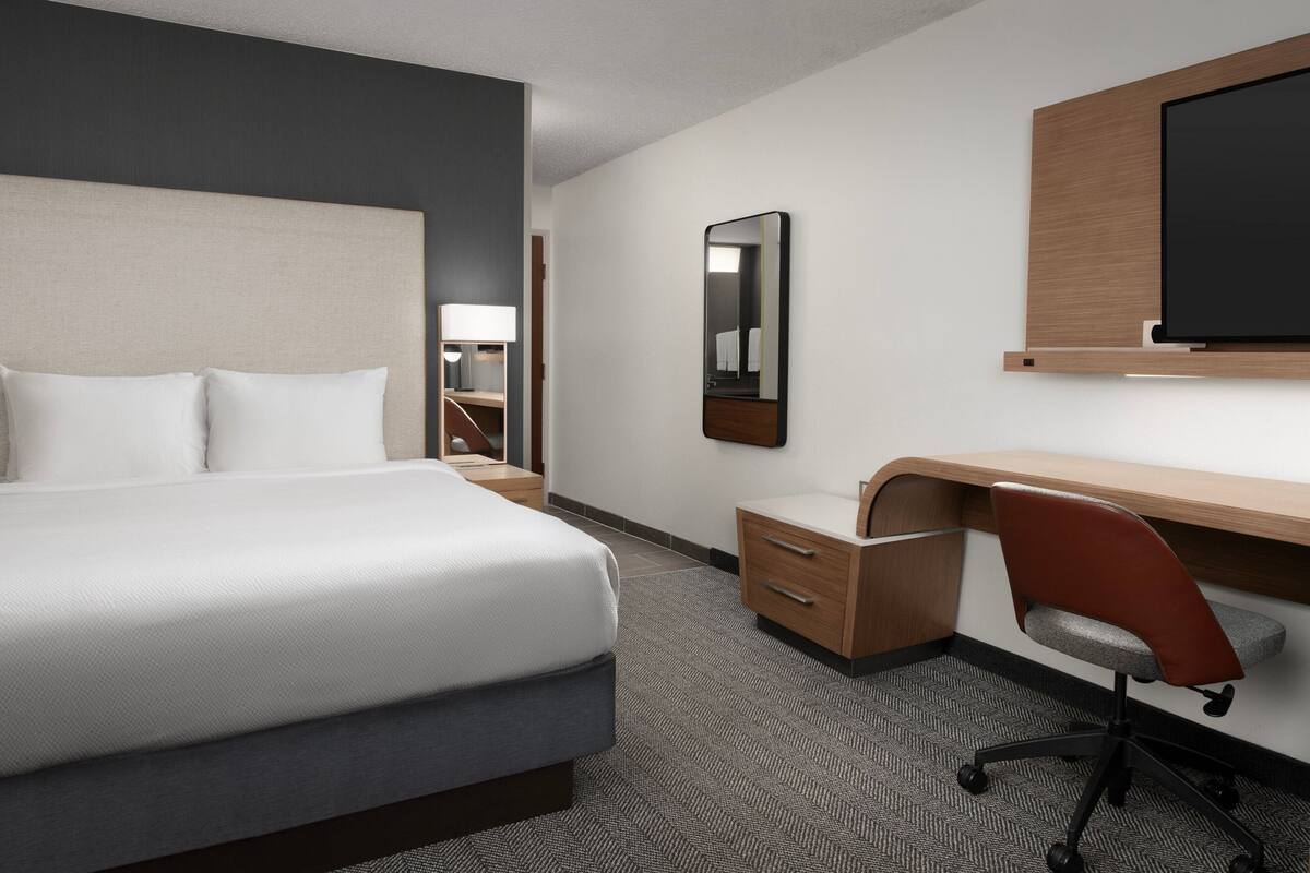 A hotel room with a bed, a desk, a chair, a mirror, and a TV.