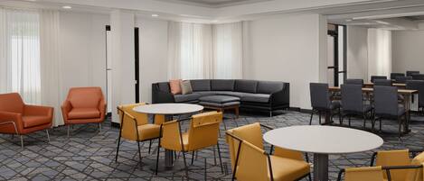 Una habitación de hotel moderna con un sofá gris, sillones naranjas, sillas amarillas y una mesa redonda.