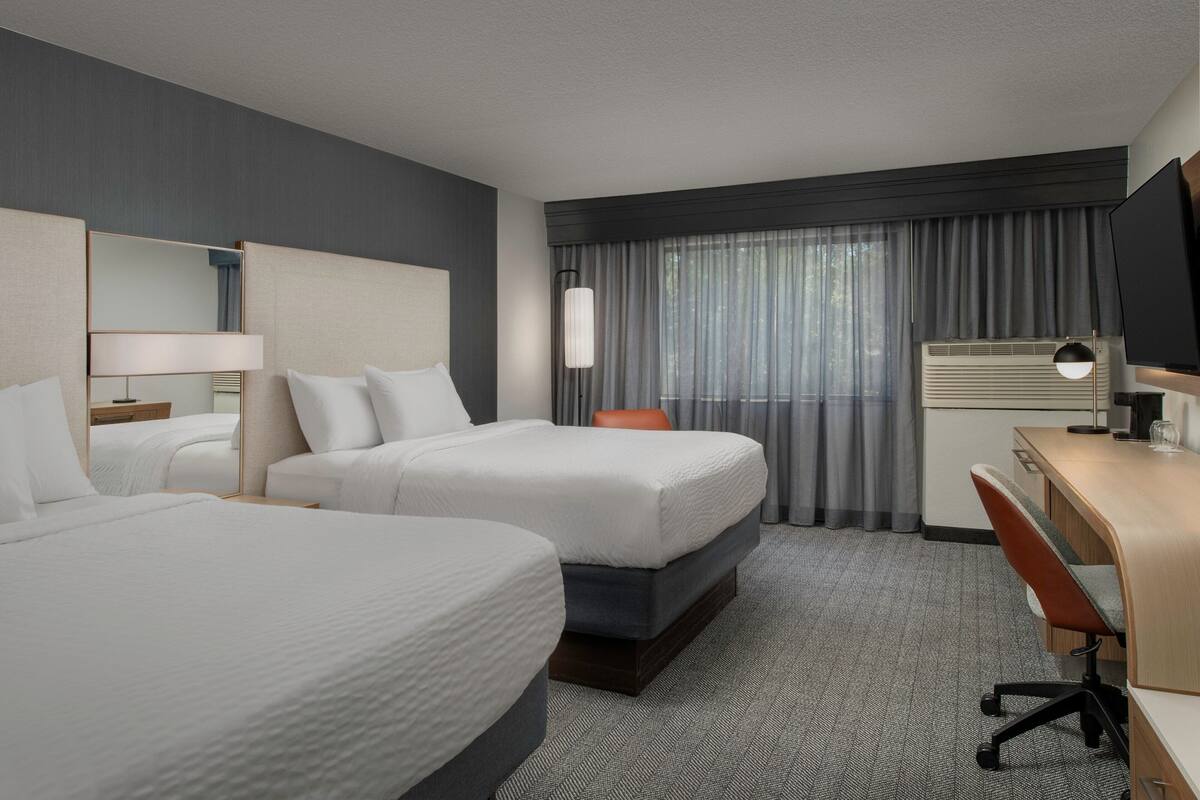 A hotel room with two beds, a desk, a chair, a TV, and a window with curtains.