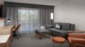 A modern hotel room with a desk, chair, sofa, ottoman, and a TV mounted on the wall. - Courtyard by Marriott Dothan (Dothan)