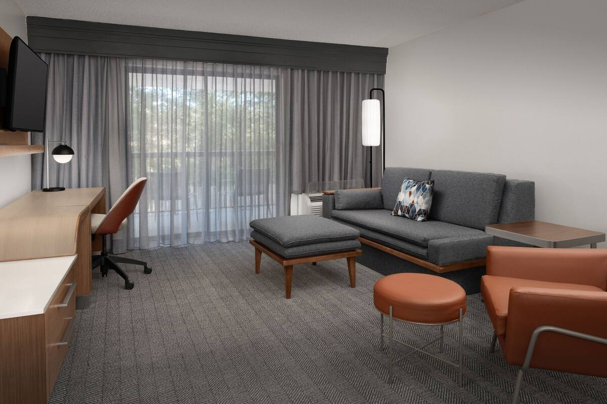 A modern hotel room with a desk, chair, sofa, ottoman, and a TV mounted on the wall.