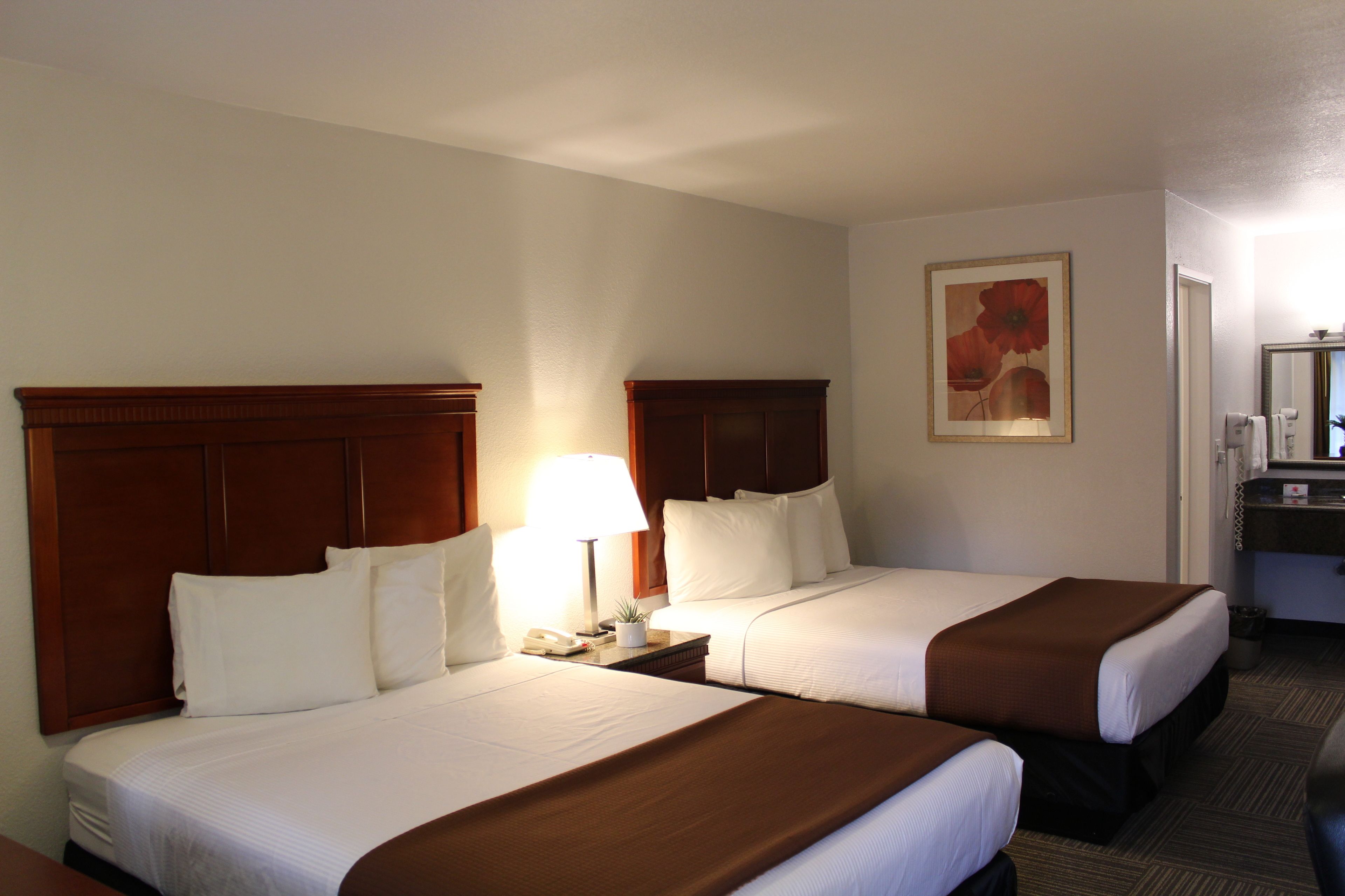 A hotel room with two beds, a nightstand with a lamp, a painting on the wall, and a bathroom visible through an open door.