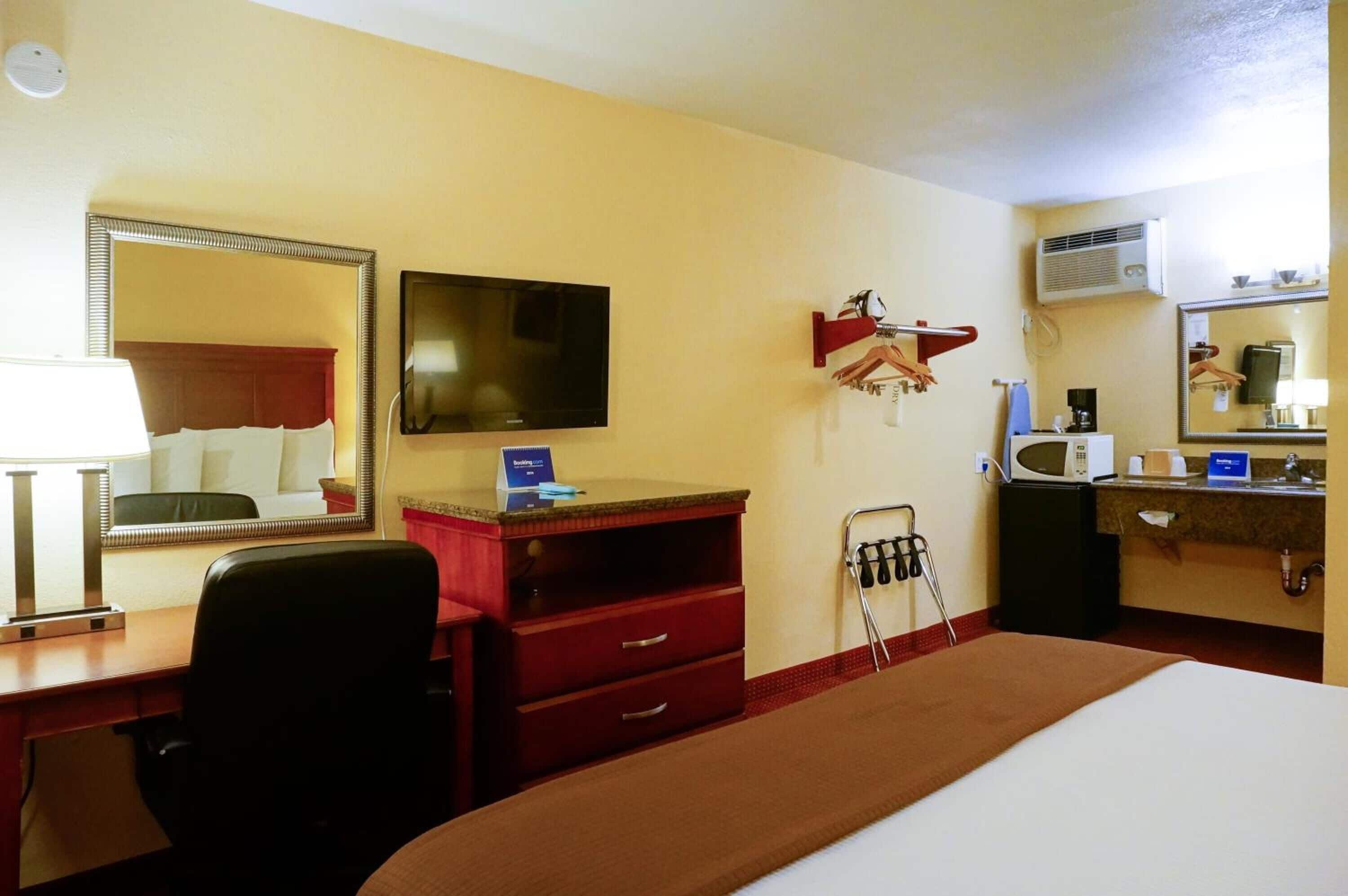 A hotel room with a bed, a desk with a computer, a TV, a mirror, a red dresser, a microwave, and a small kitchenette.
