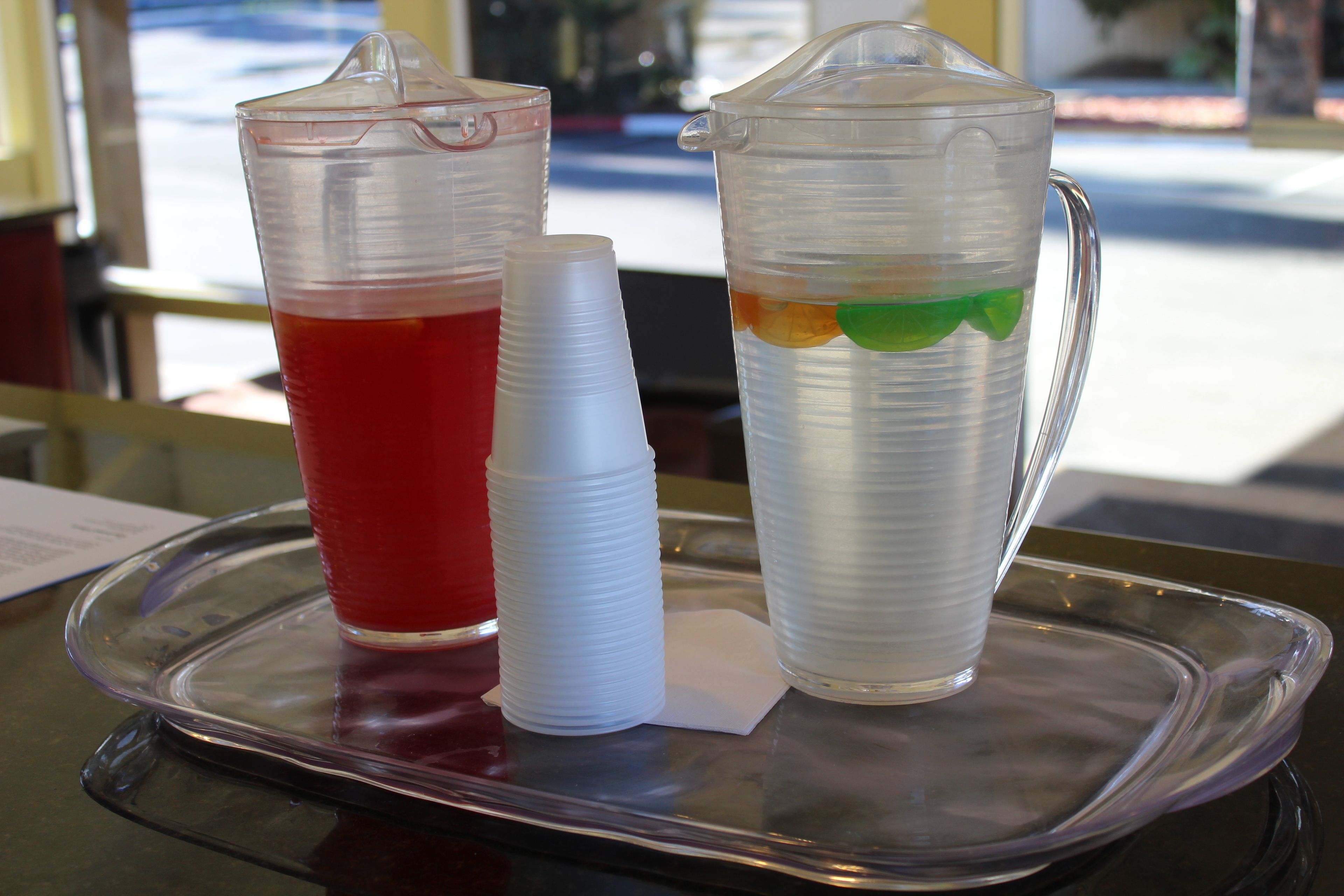 Un vassoio con due bicchieri di plastica e una brocca, uno pieno di liquido rosso e l'altro di liquido trasparente con un oggetto verde.