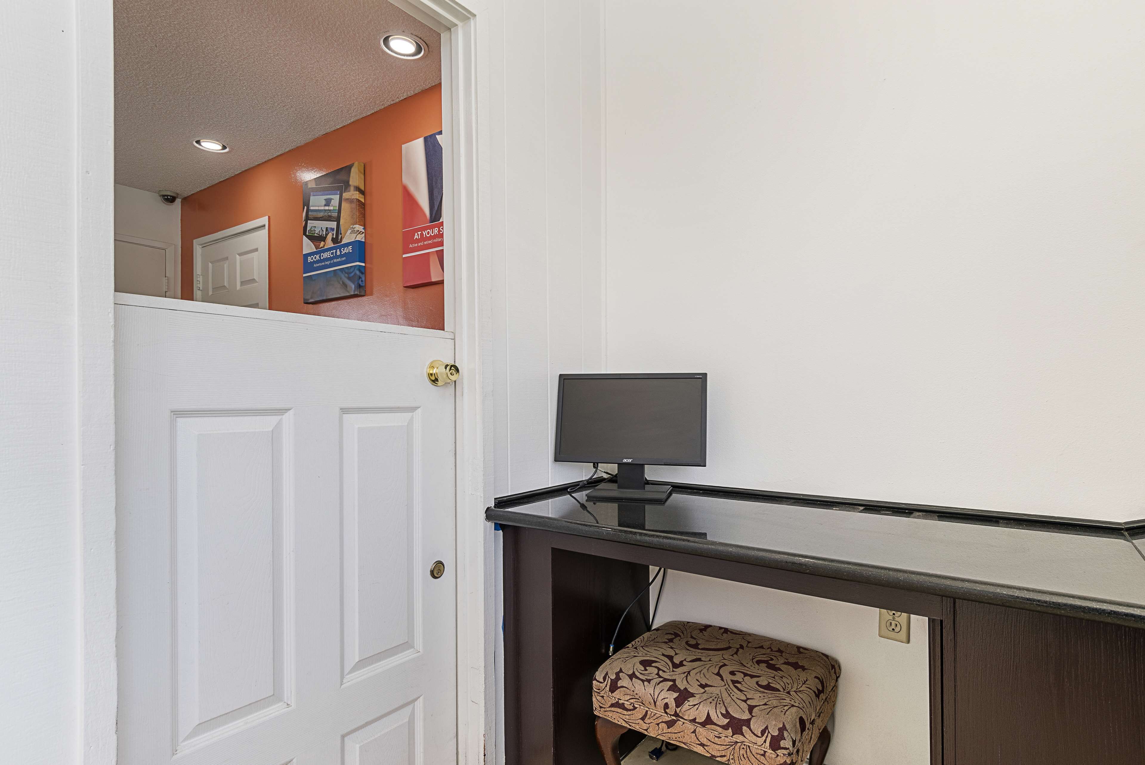 A room with a white door, a small desk with a monitor, and a patterned ottoman.