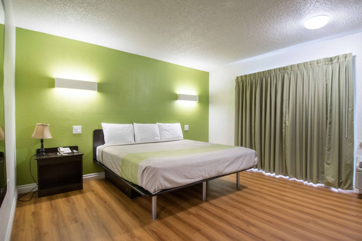 A hotel room with a bed, green walls, a nightstand with a lamp, and a window with curtains.