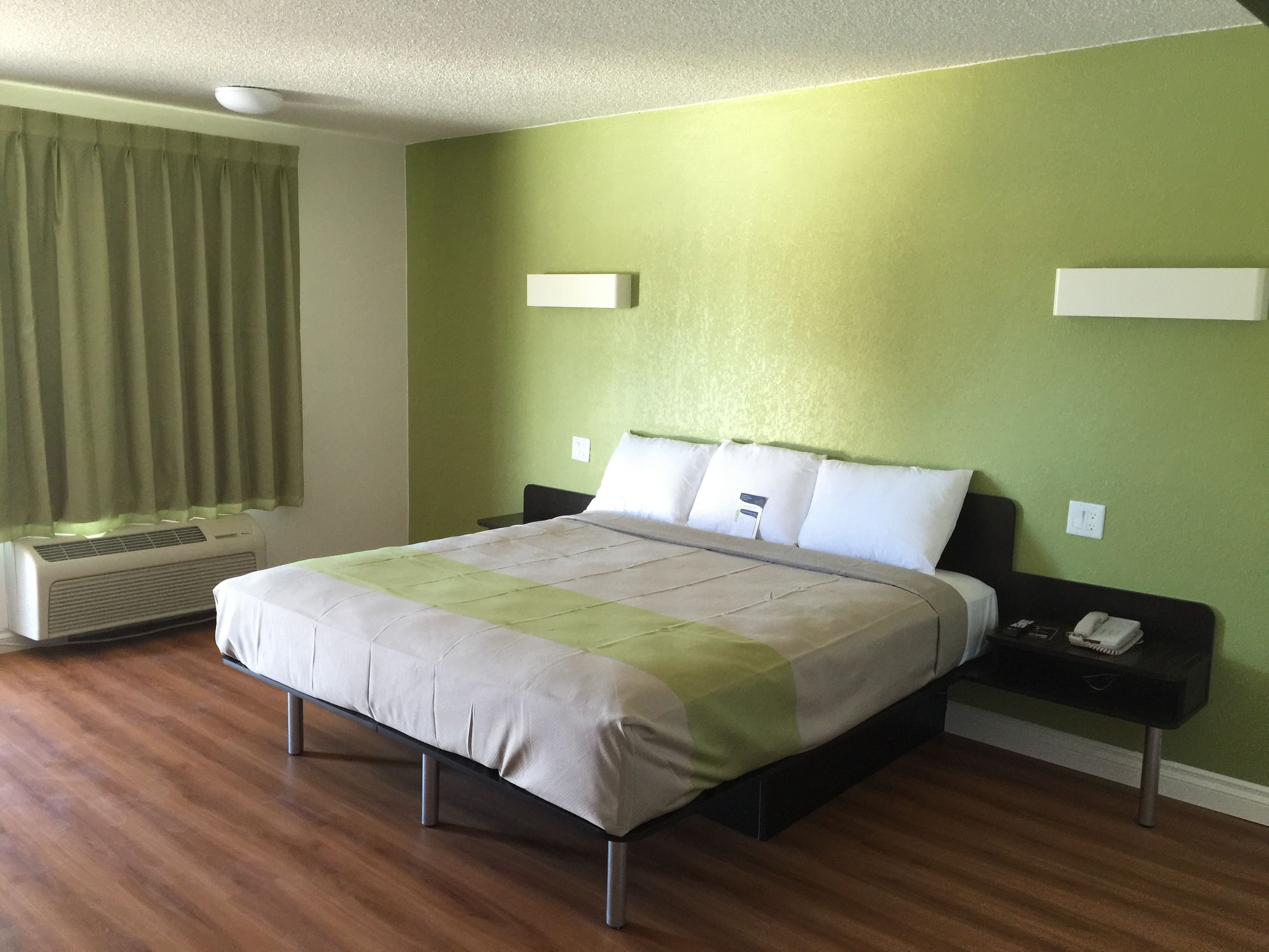 A neatly made bed with a green and white bedspread in a room with green walls, a wooden floor, and a window with curtains.