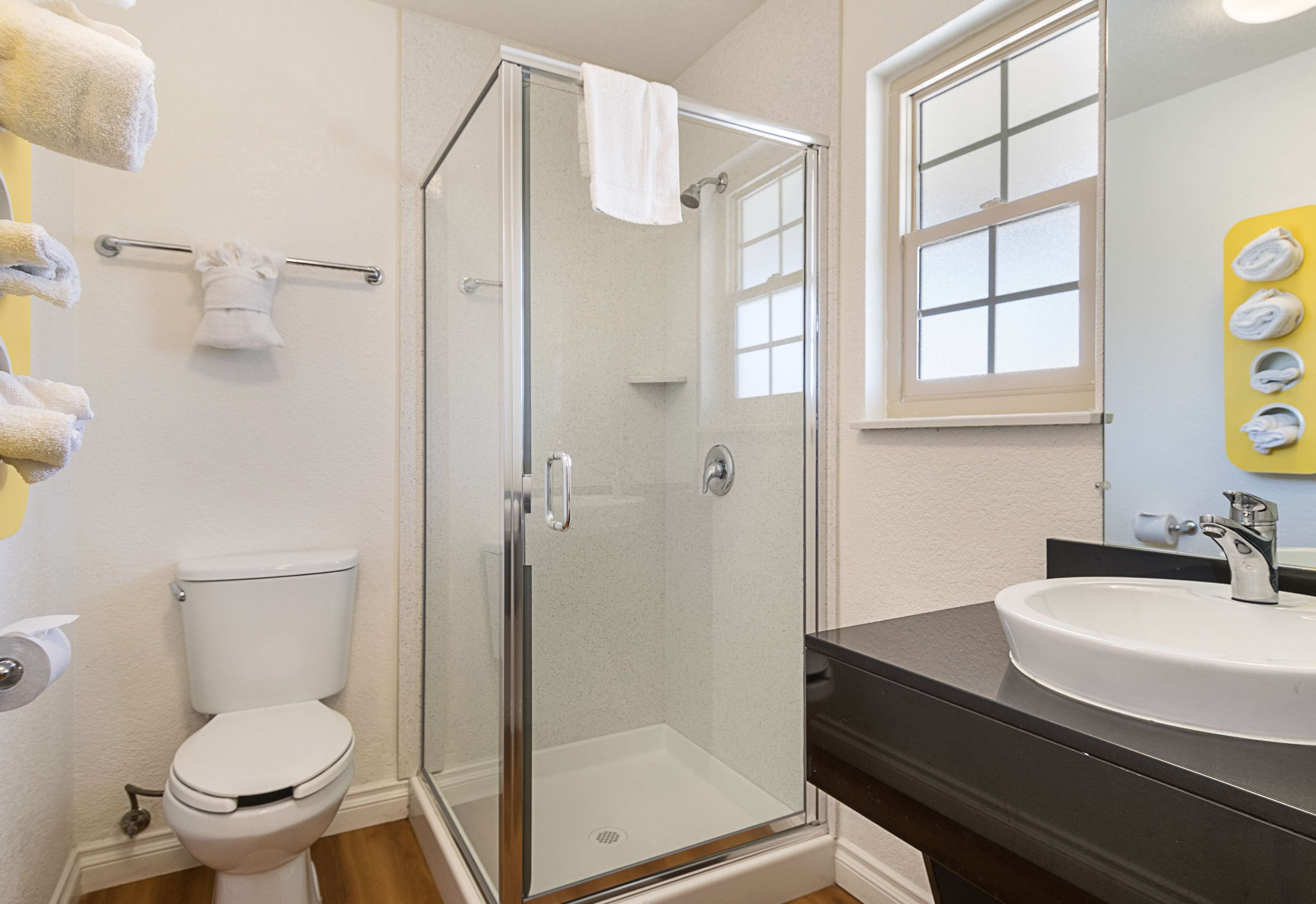 A bathroom with a glass-enclosed shower, a white toilet, a sink with a black countertop, and a mirror with a window above it.